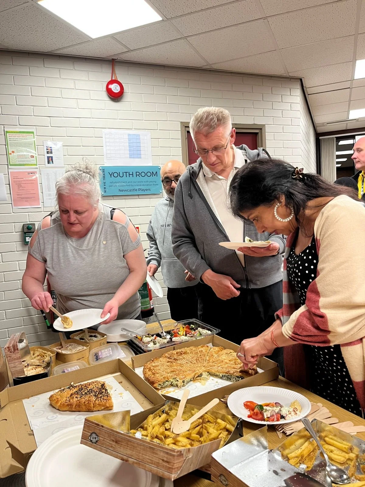 3 adults stand around a table in a white room, helping themselves to a buffet of pizza and potato wedges