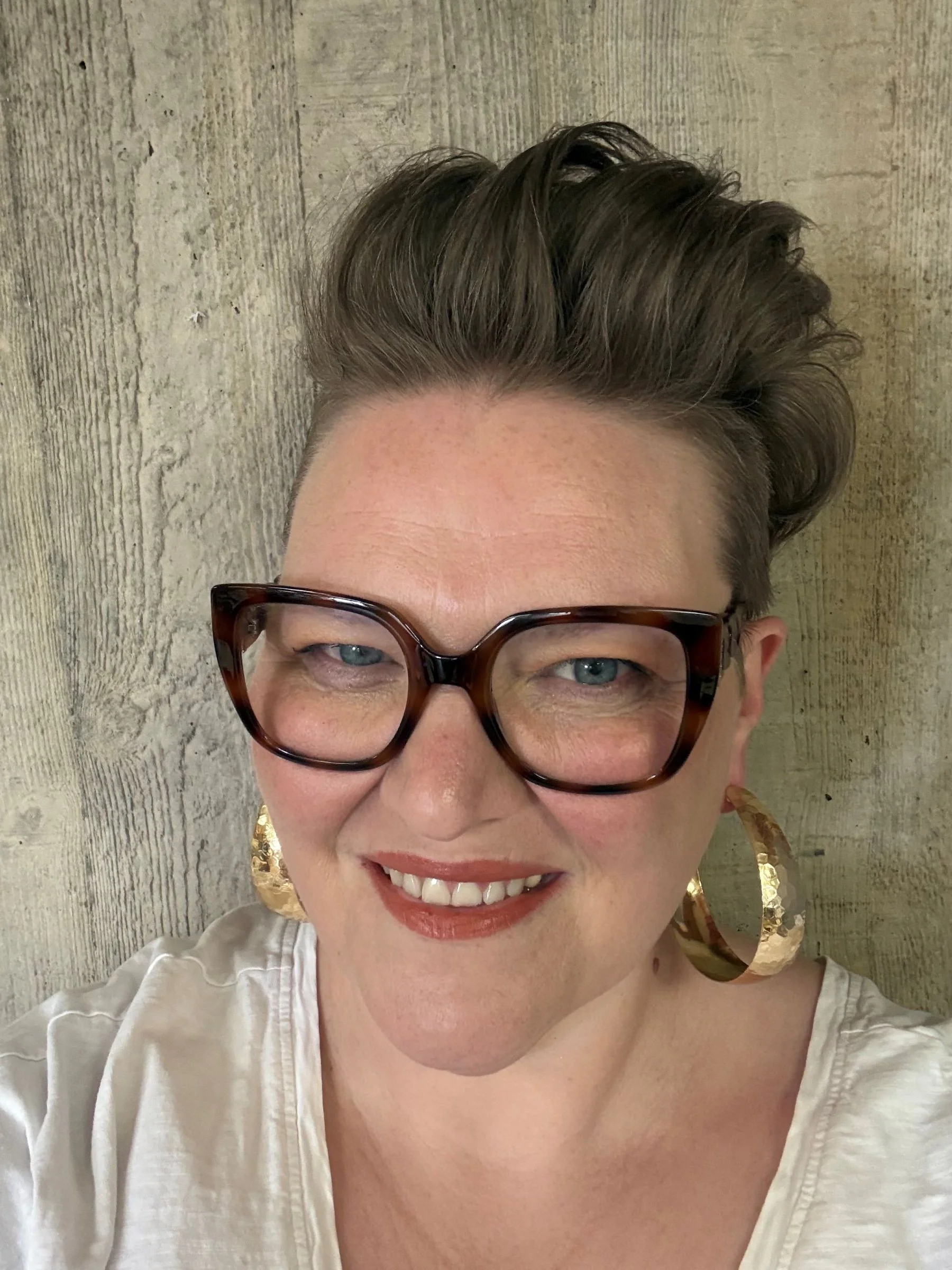 Head and shoulder shot of Mel, a white, middle-aged woman with high pompadour hair shaved around the sides. She's wearing a white T-shirt, large gold earrings, and improbably massive tortoise shell glasses.