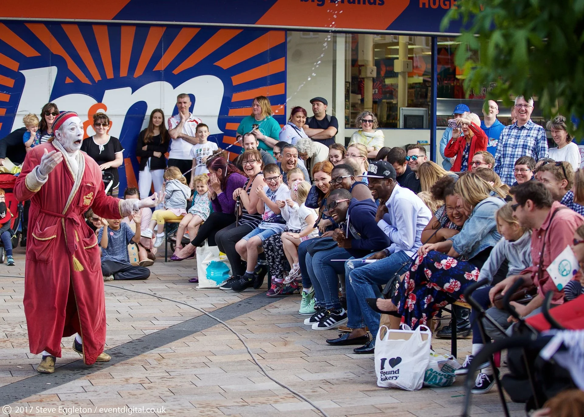 A street performer dressed as a clown with ghostly makeup and red and beige robes performing for an audience in a public space, with a large crowd of smiling and laughing people of various ages watching.