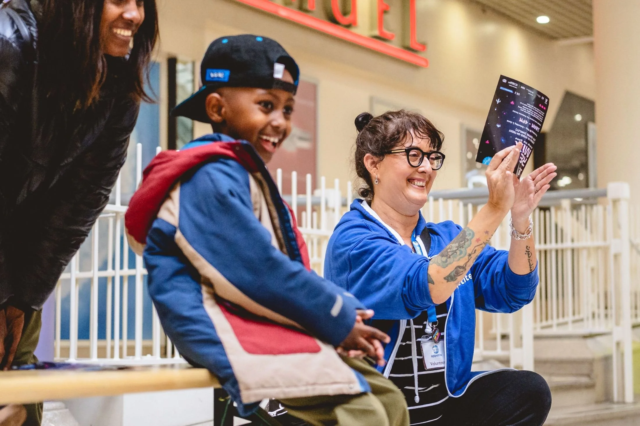In the background, a volunteer in blue hoodie is smiling and clapping, holding an event flyer. In the foreground is a Black child in a blue coat and black cap with a big smile.