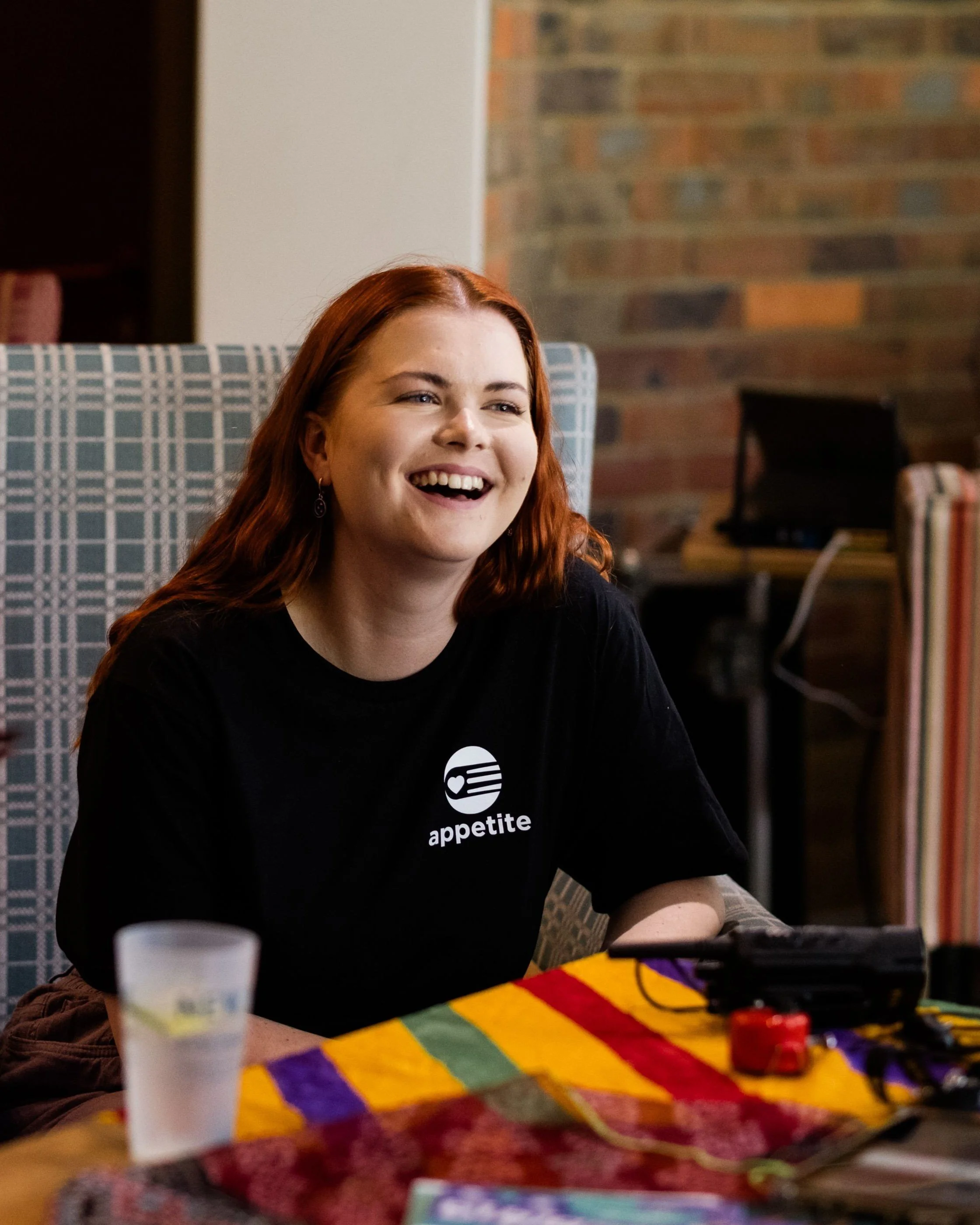 A white woman with long auburn hair wearing a black T-shirt with the appetite logo sits in a chair with a laughing smile.
