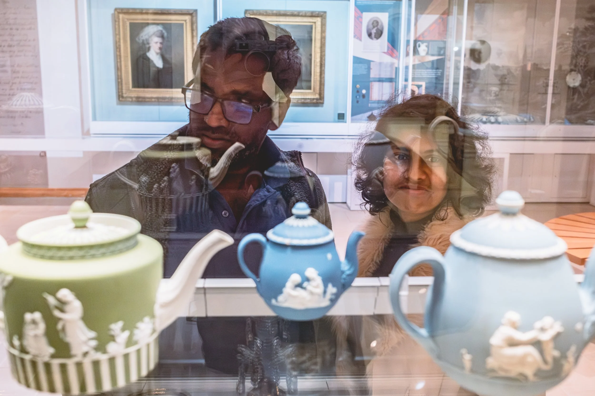 A smiling Asian woman and an Asian man in glasses look through a glass cabinet in a gallery at Wedgwood ceramics. A green teapot is on the left, a light blue teapot is on the right and a smaller, blue teapot in the middle.