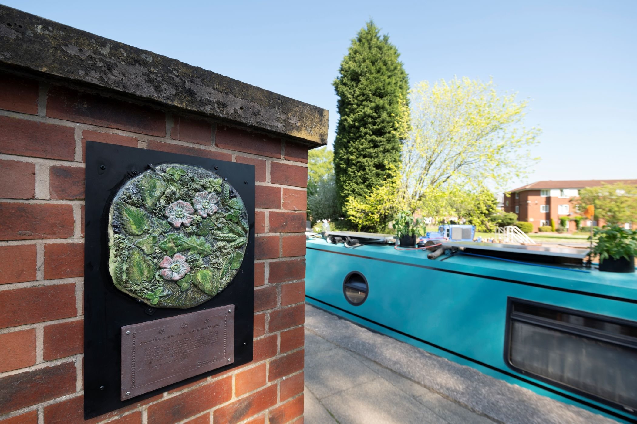 A close up of a circular ceramic plaque on a brick wall featuring green leaves and white flowers. A light blue canal boat is in the background.