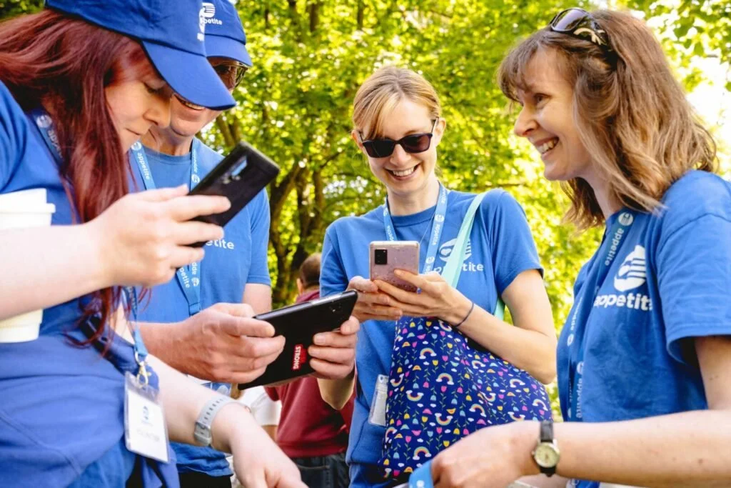A side-on view of Heather wearing a blue Appetite T-shirt. She has shoulder-length brown hair with large brown sunglasses pushed on top of her head holding the hair back. Heather is outside on a sunny day, smiling and with a group of volunteers.