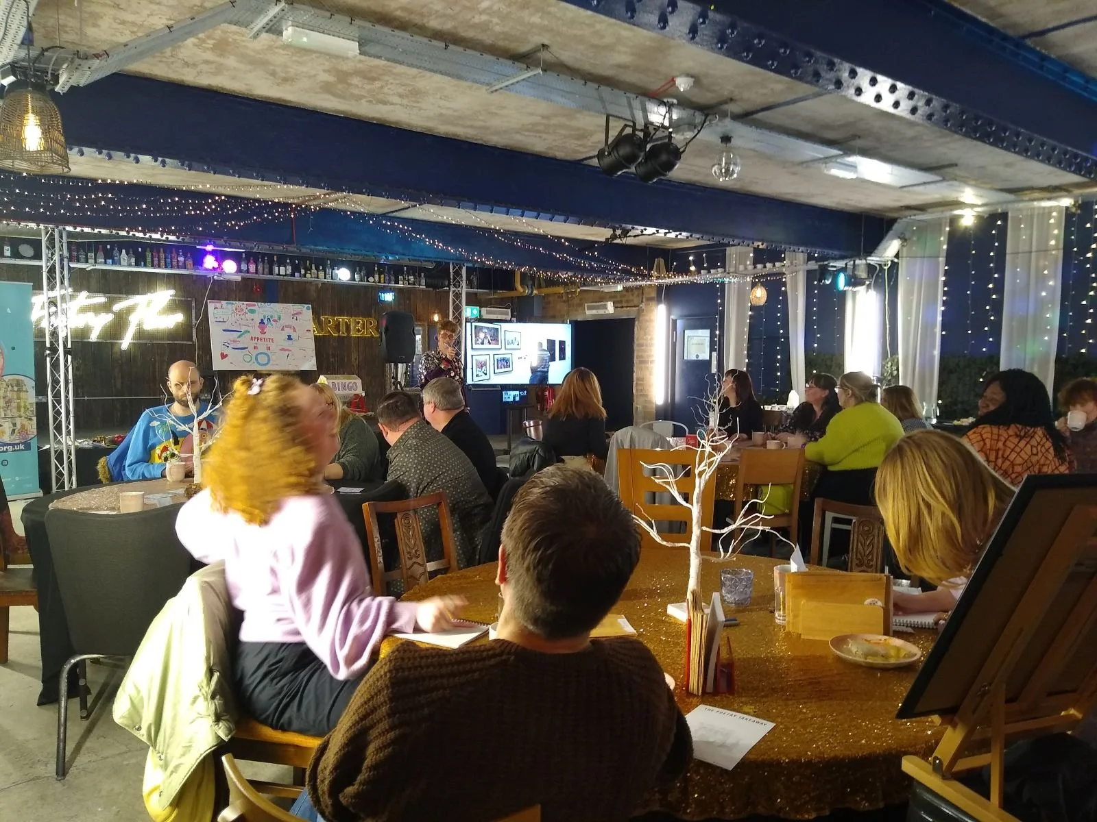A wide image of adults sat around circular tables like a conference with a display screen in the background. The room has an industrial feel, painted blue and decorated with fairy lights.