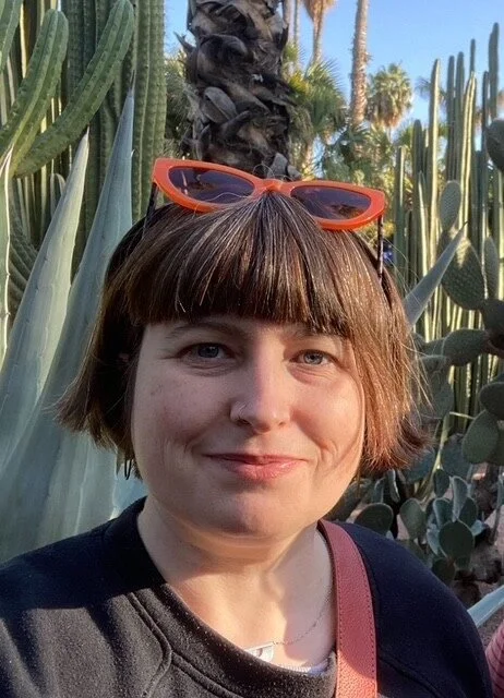 A white woman with short dark bobbed hair and a fringe is smiling into the camera. She has orange sunglasses peached on her head and a sliver nose hoop. She is wearing a black jumper and there are large cacti in the background.