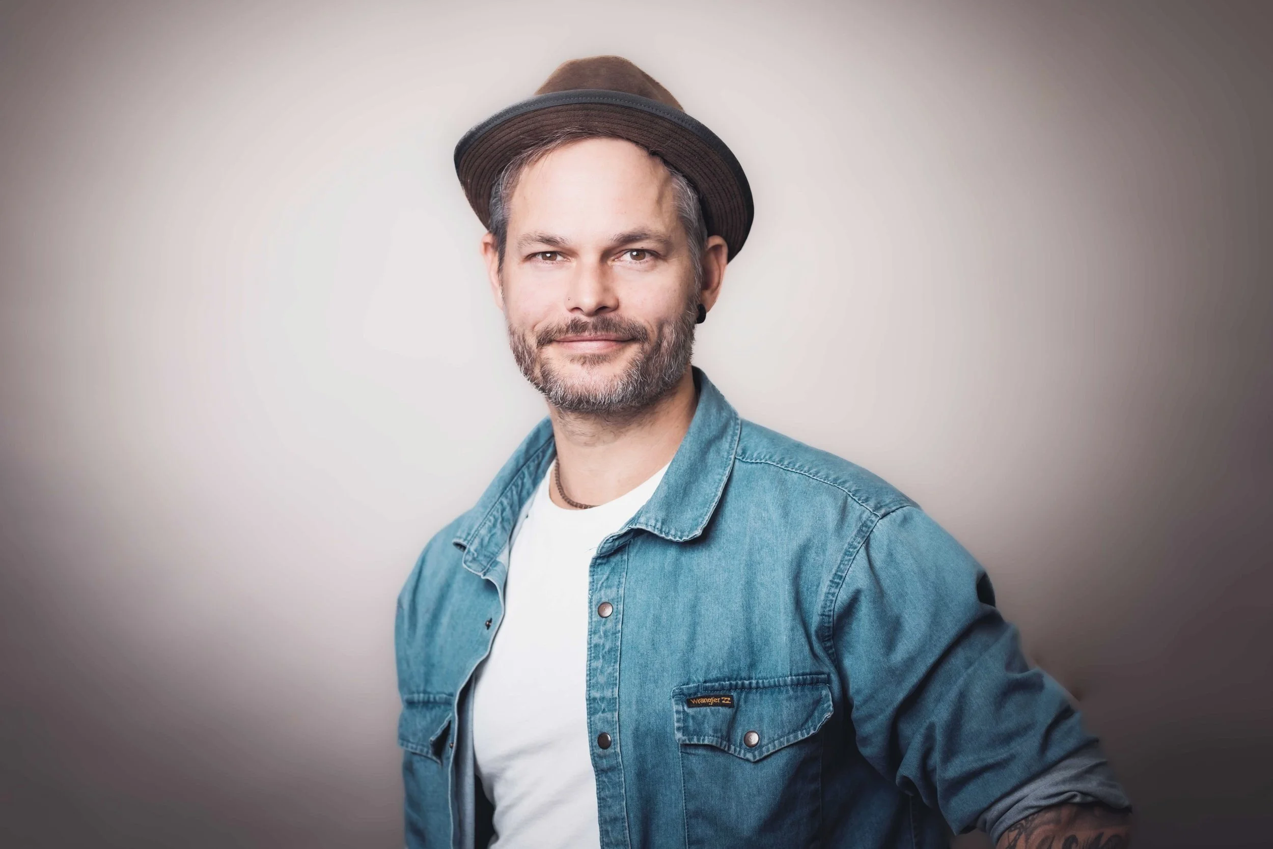 Mann mit Hut, blauer Jeansjacke, weißen T-Shirt posiert vor grauem Hintergrund.