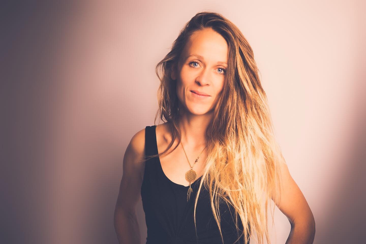 Junge Frau mit longem, lockigem, blondem Haar trägt schwarze Top und eine goldene Halskette steht vor hellem, neutrales Hintergrund.