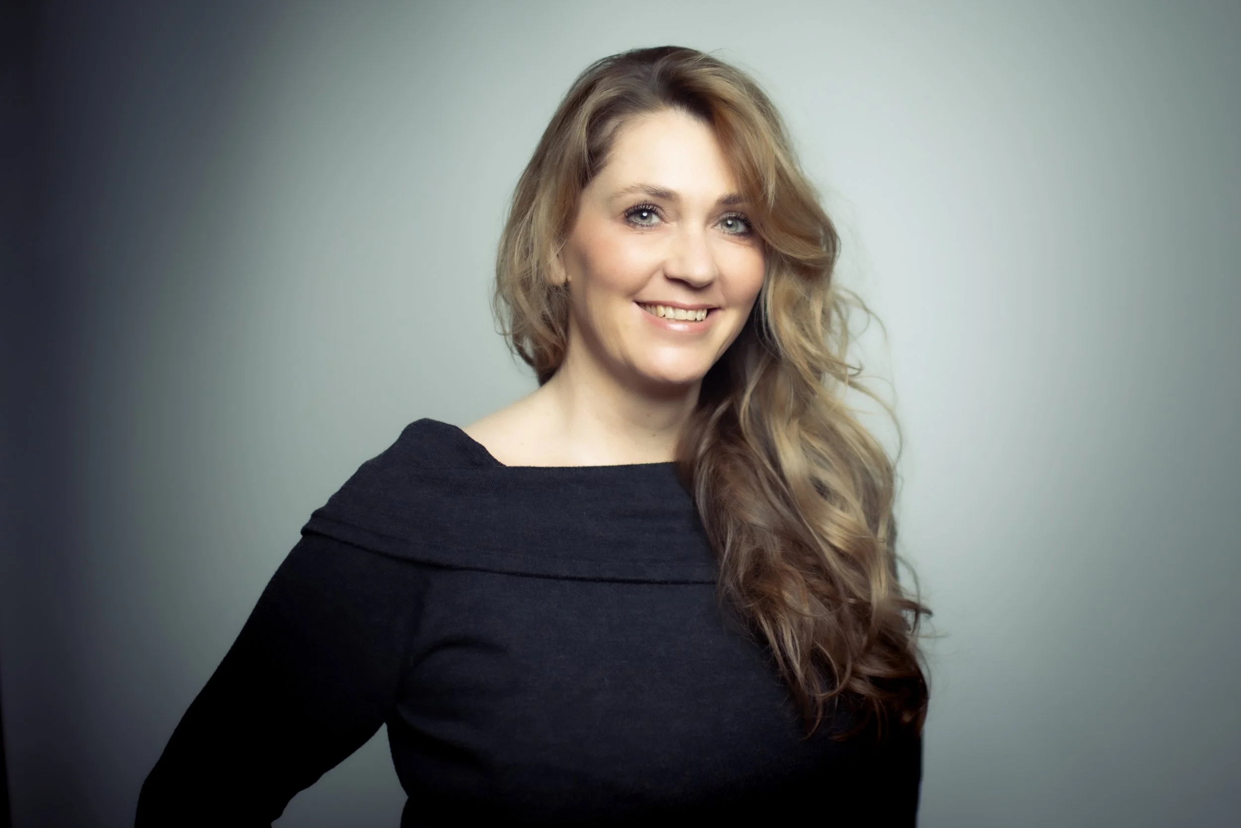 Eine lächelnde Frau mit langem, gewelltem, hellbraunem Haar in einem schwarzen Oberteil vor einem grauen Hintergrund.