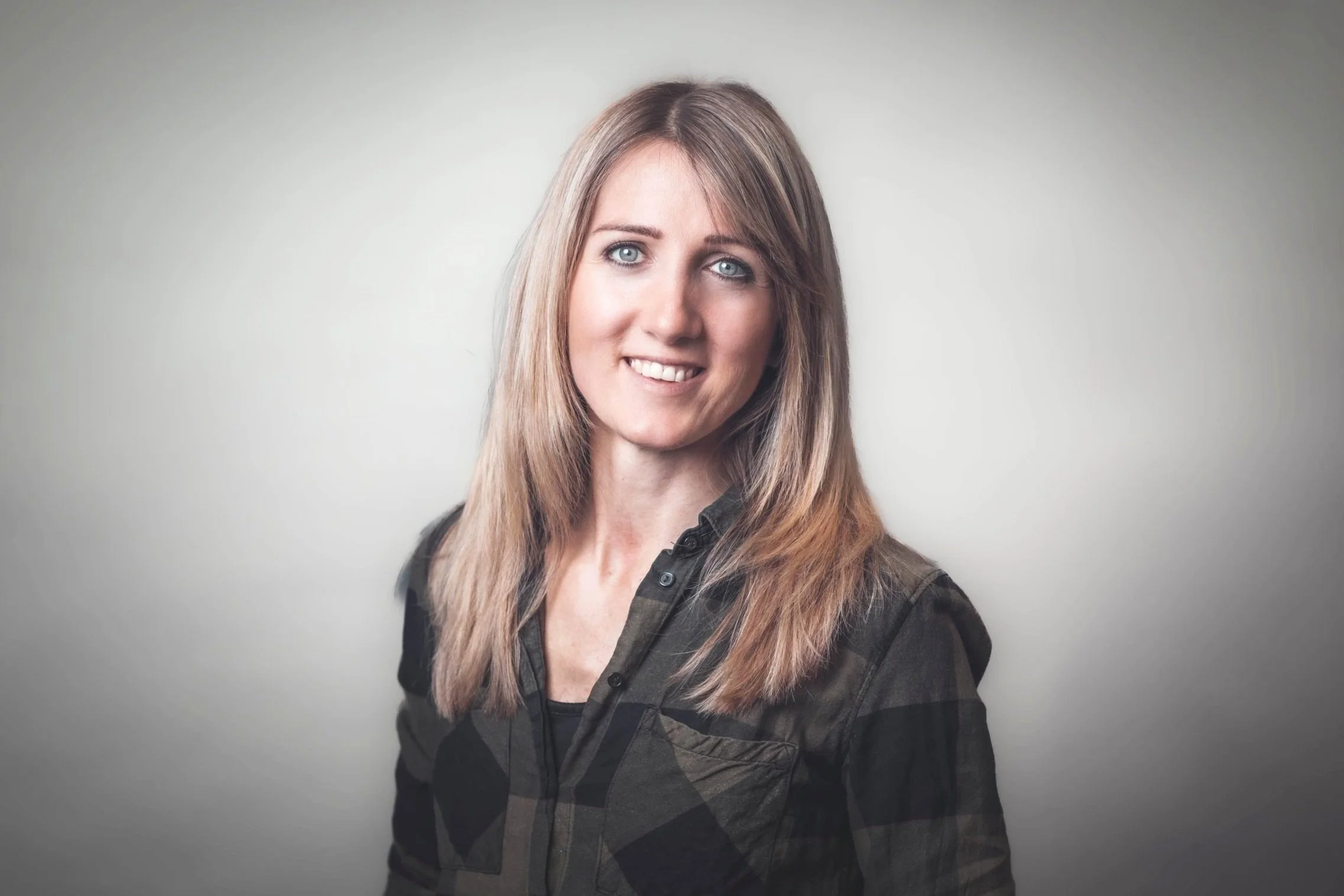 Junge Frau mit braunen Haaren, blauen Augen und freundlichem Lächeln, trägt eine dunkle kariert Jacke, vor hellem Hintergrund.