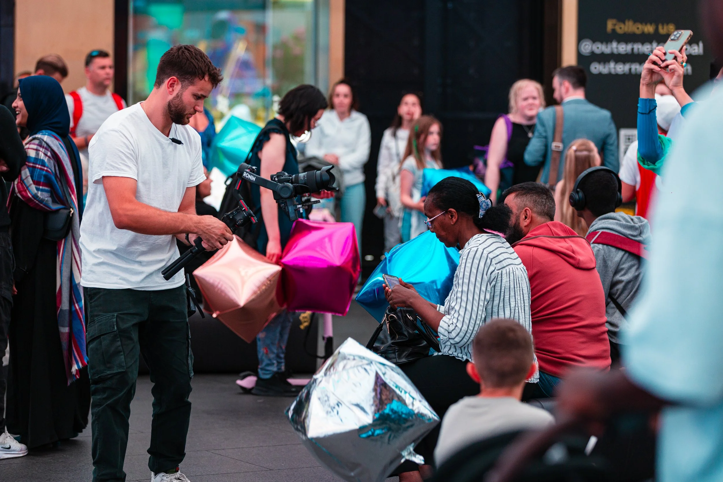 A man filming with a camera on a stabilizer in a crowded outdoor setting filled with people, some sitting and some standing, holding colorful balloons and bags, with a black wall and signage in the background.