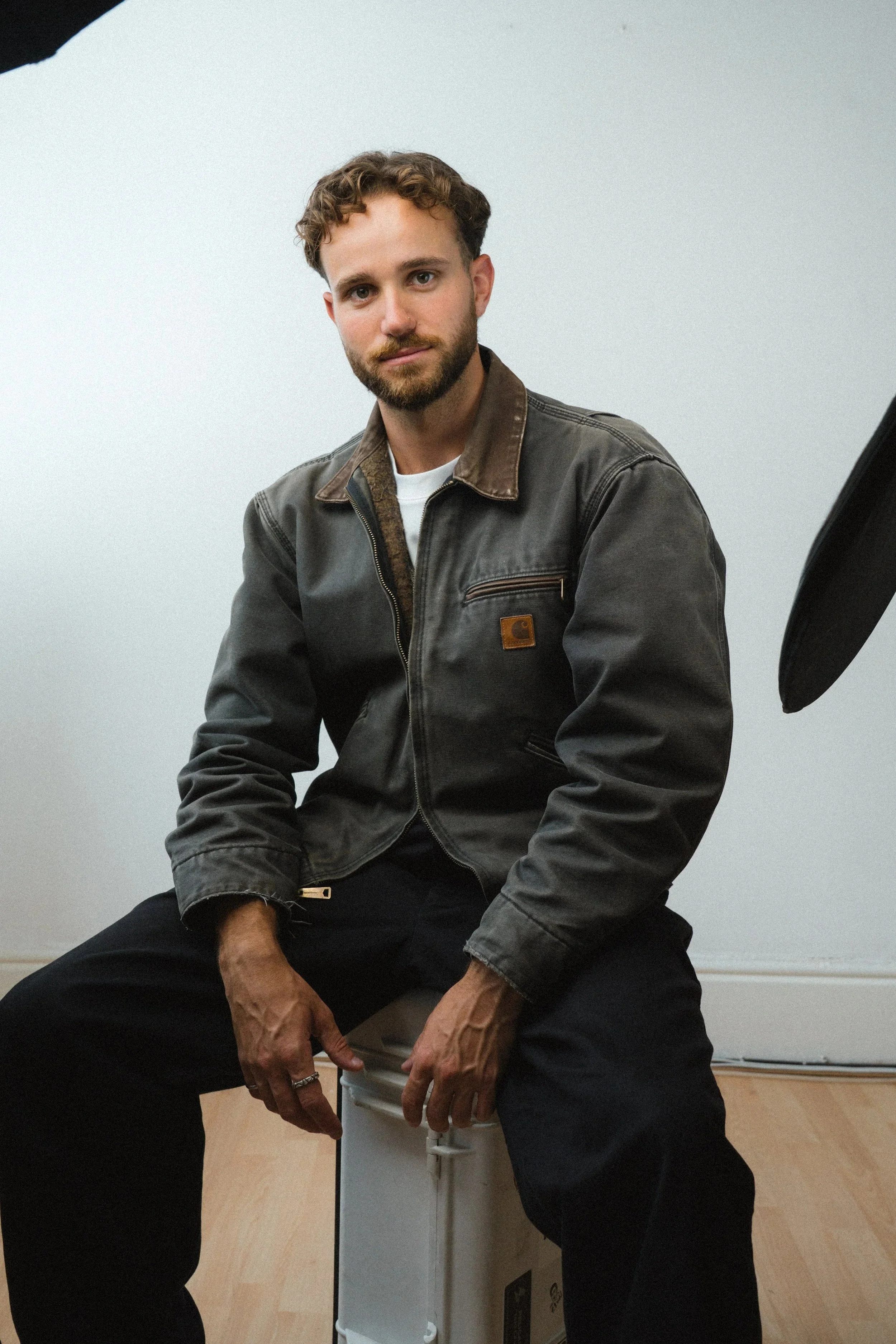 A man with curly brown hair and a beard sitting on a white box against a plain white wall, wearing a black leather jacket, white shirt, and black pants.