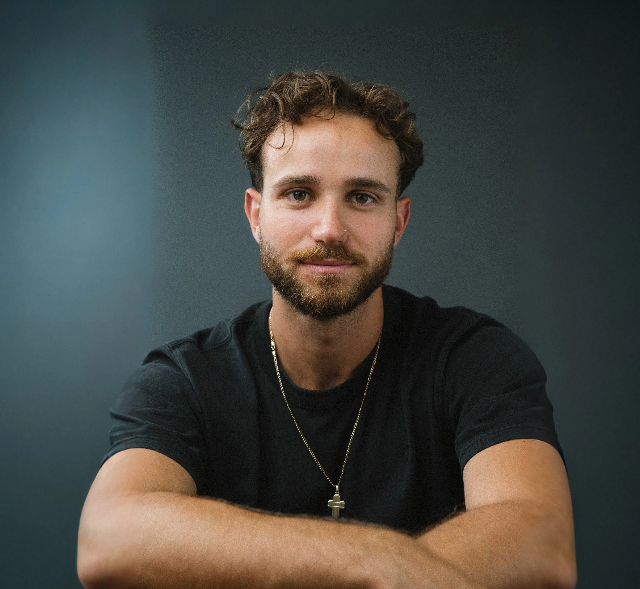 Portrait of a young man with brown curly hair and a beard, wearing a black t-shirt and a cross necklace, against a dark green background.