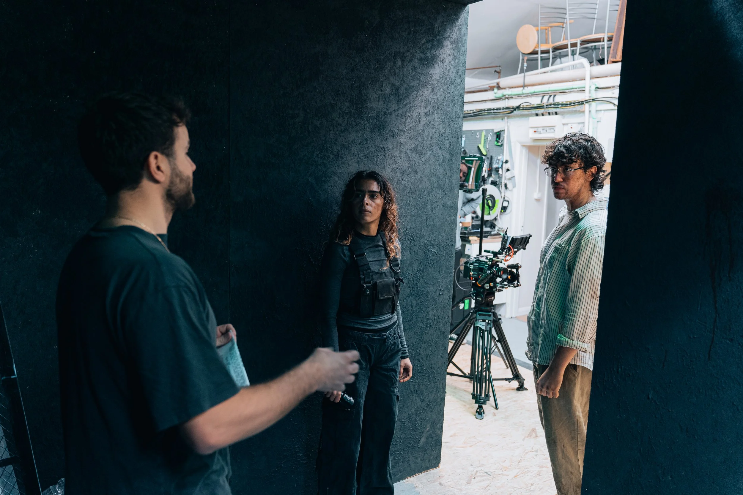 Three people on a film set, two men and a woman, with camera equipment and lighting in the background.