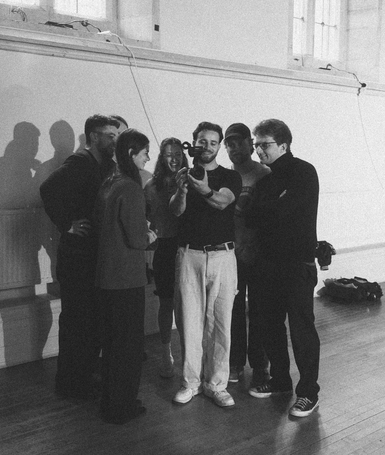 Group of six young adults taking a selfie in black and white, standing indoors against a plain wall, with some shadows cast on the wall.