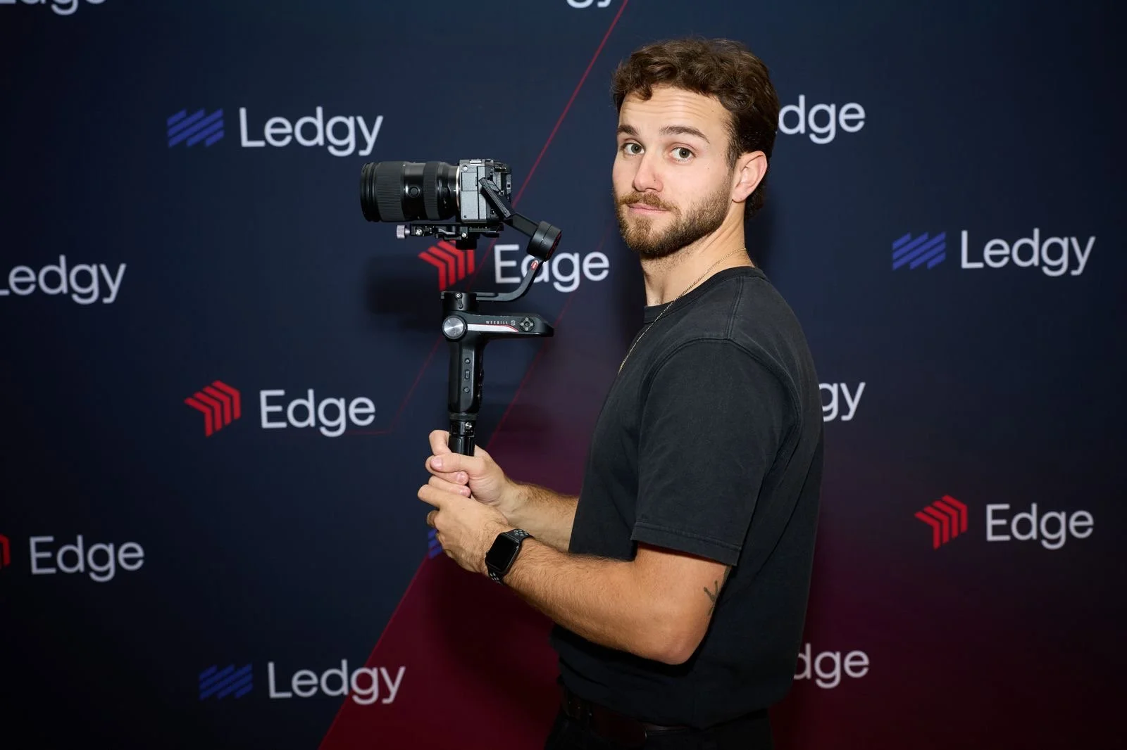 A young man with brown hair and a beard, wearing a black shirt, holding a camera stabilizer with a camera mounted on it, standing in front of a black background with the words 'Ledgy' and 'Edge' printed repeatedly.