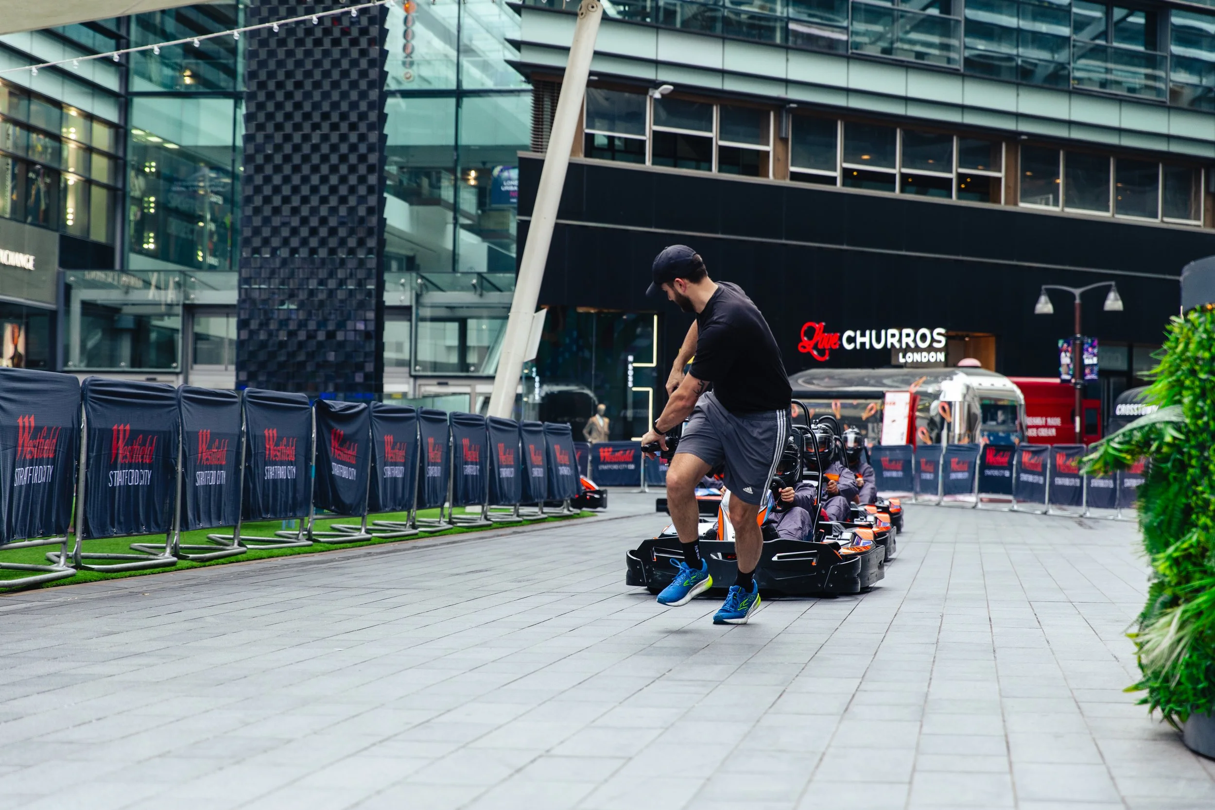 A man riding a go-kart on an outdoor city street, with a modern glass building and a churros stand in the background.
