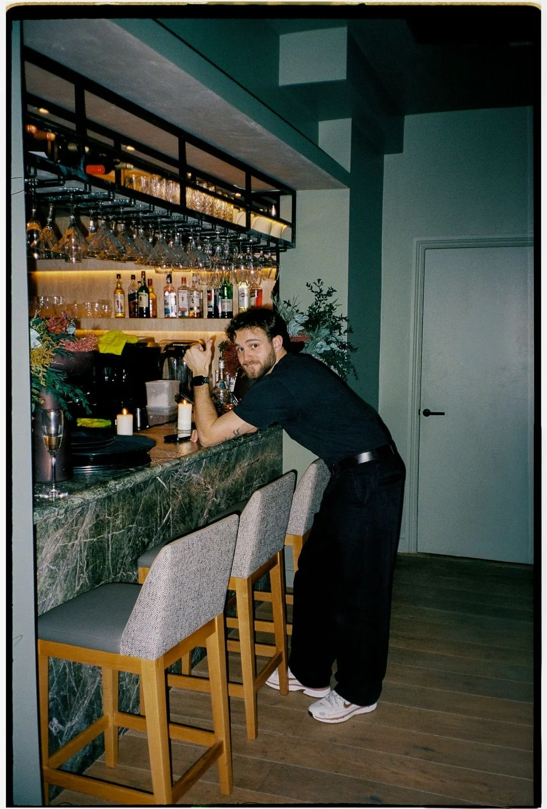 A man leaning on a bar counter, smiling, with bar shelves filled with liquor bottles and glassware behind him. Two bar stools are in front of the counter, and there are candles and plants nearby.