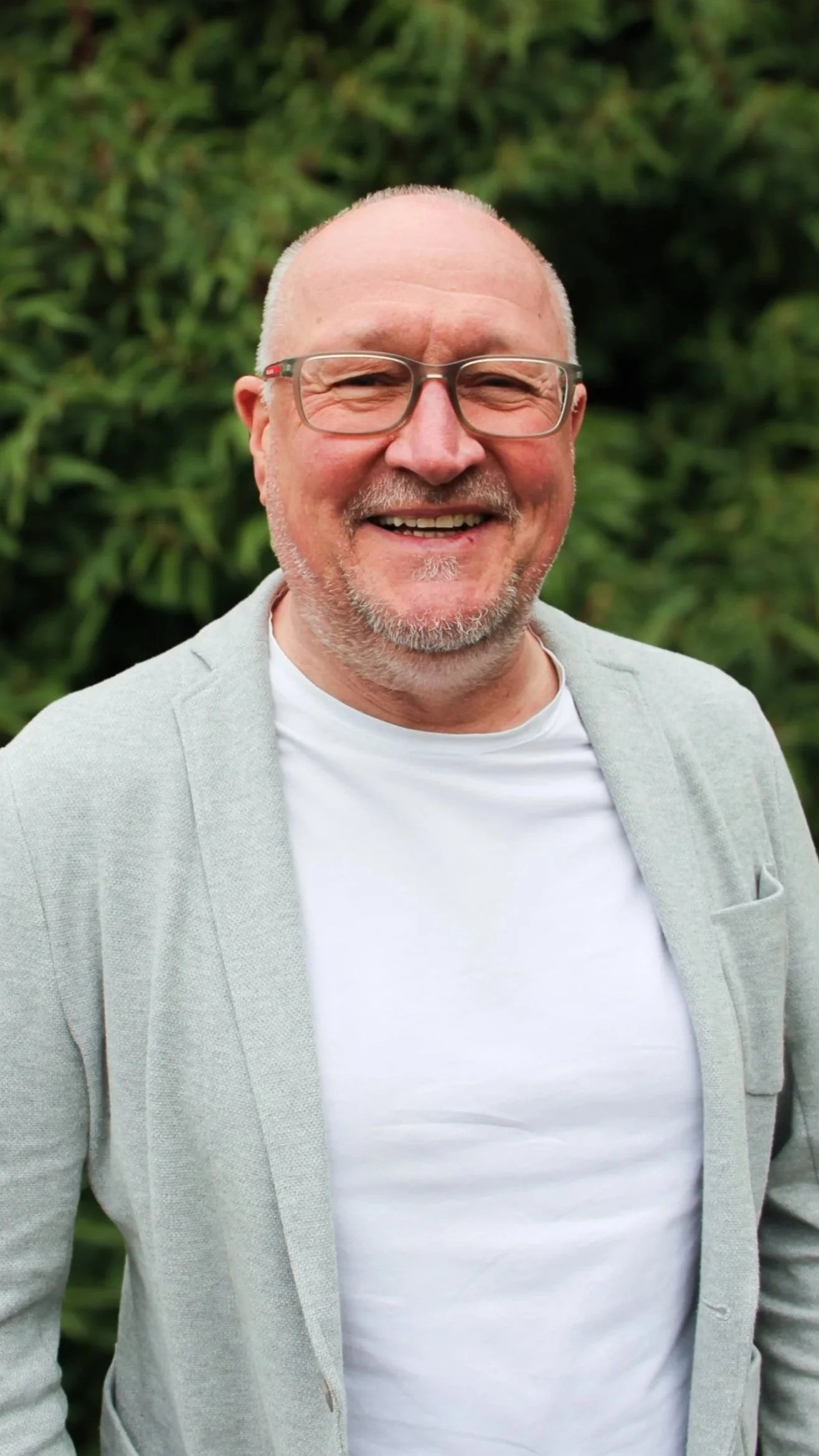 Ein lächelnder Mann mit Brille, grauem Bart und Glatze, trägt einen hellgrauen Jacke und ein weißes T-Shirt, vor grünem Busch