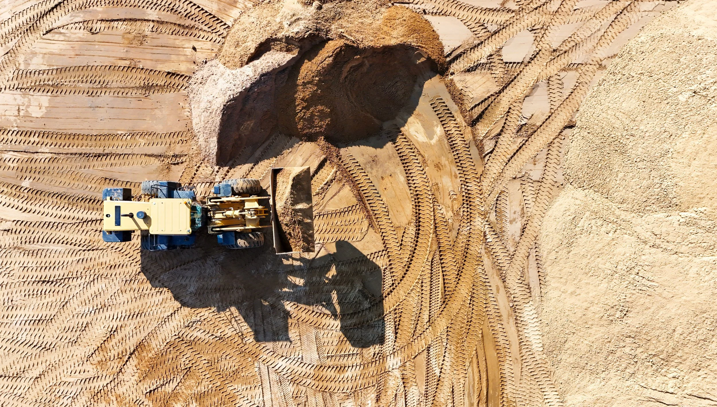 Ein Bagger arbeitet auf einer Sand- oder Kiesgrube, hinterlässt Spurenschrift im Sand, aufgenommen aus der Vogelperspektive.