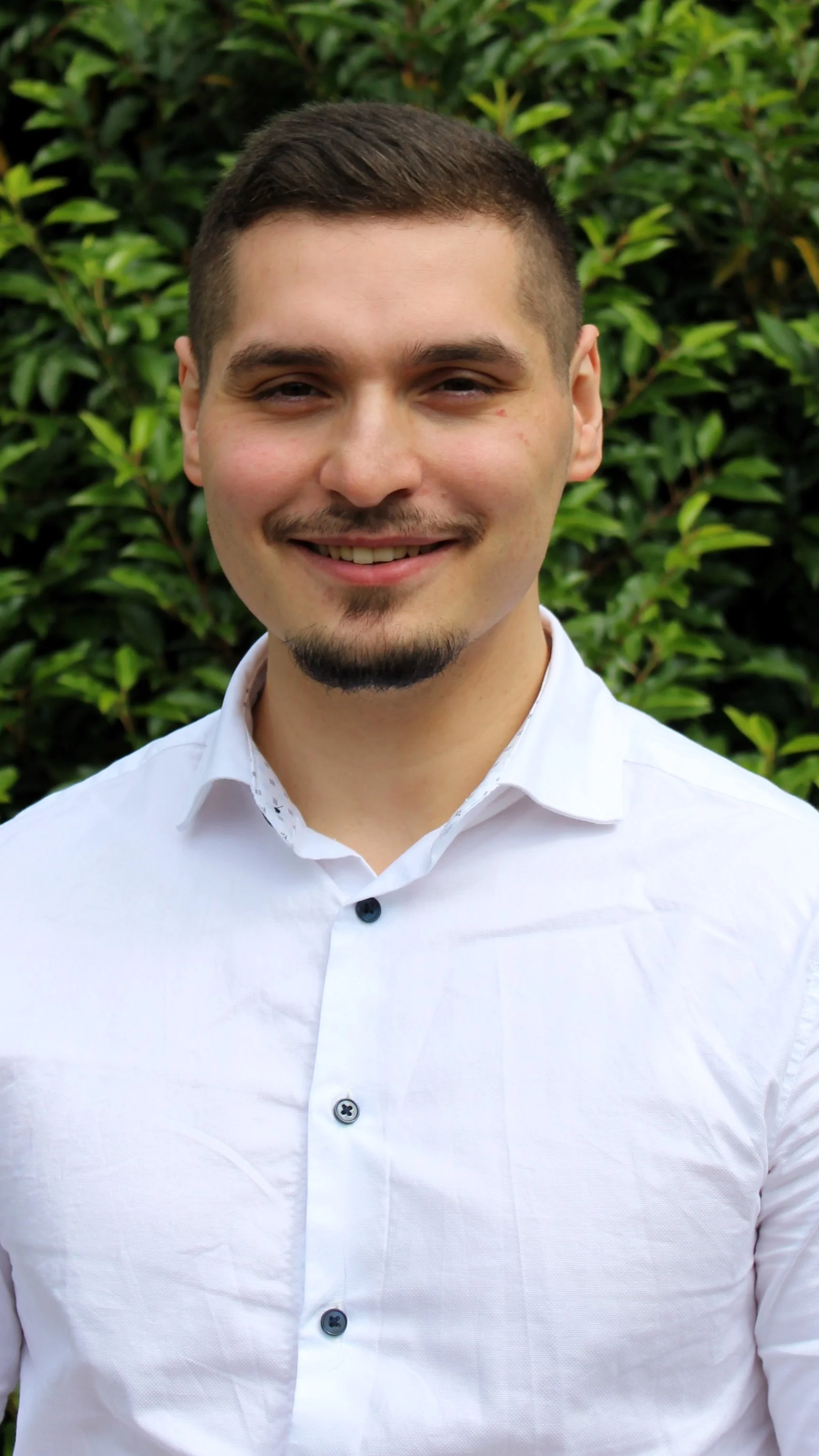 Porträt eines jungen Mannes, der in die Kamera lächelt, vor grüner Natur im Hintergrund, trägt ein weißes Hemd.