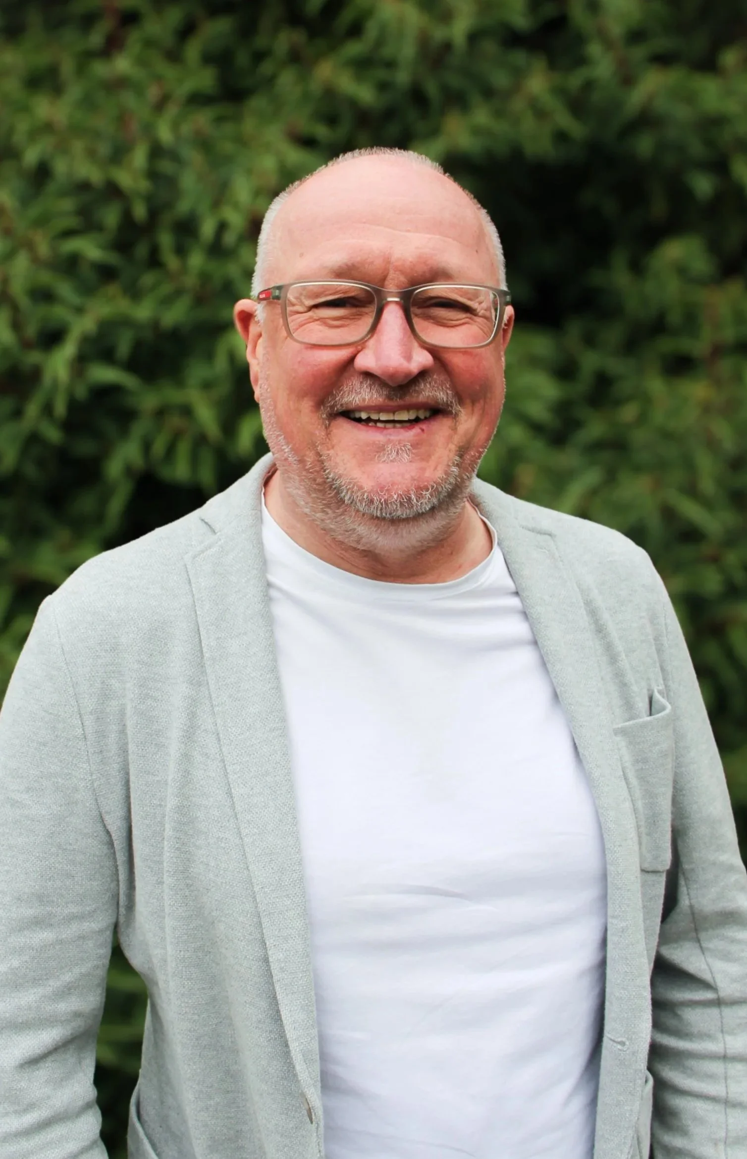 Ein lächelnder Mann mit Brille, Bart und Glatze, trägt ein hellgraues Jackett und ein weißes T-Shirt, vor grüner Natur.
