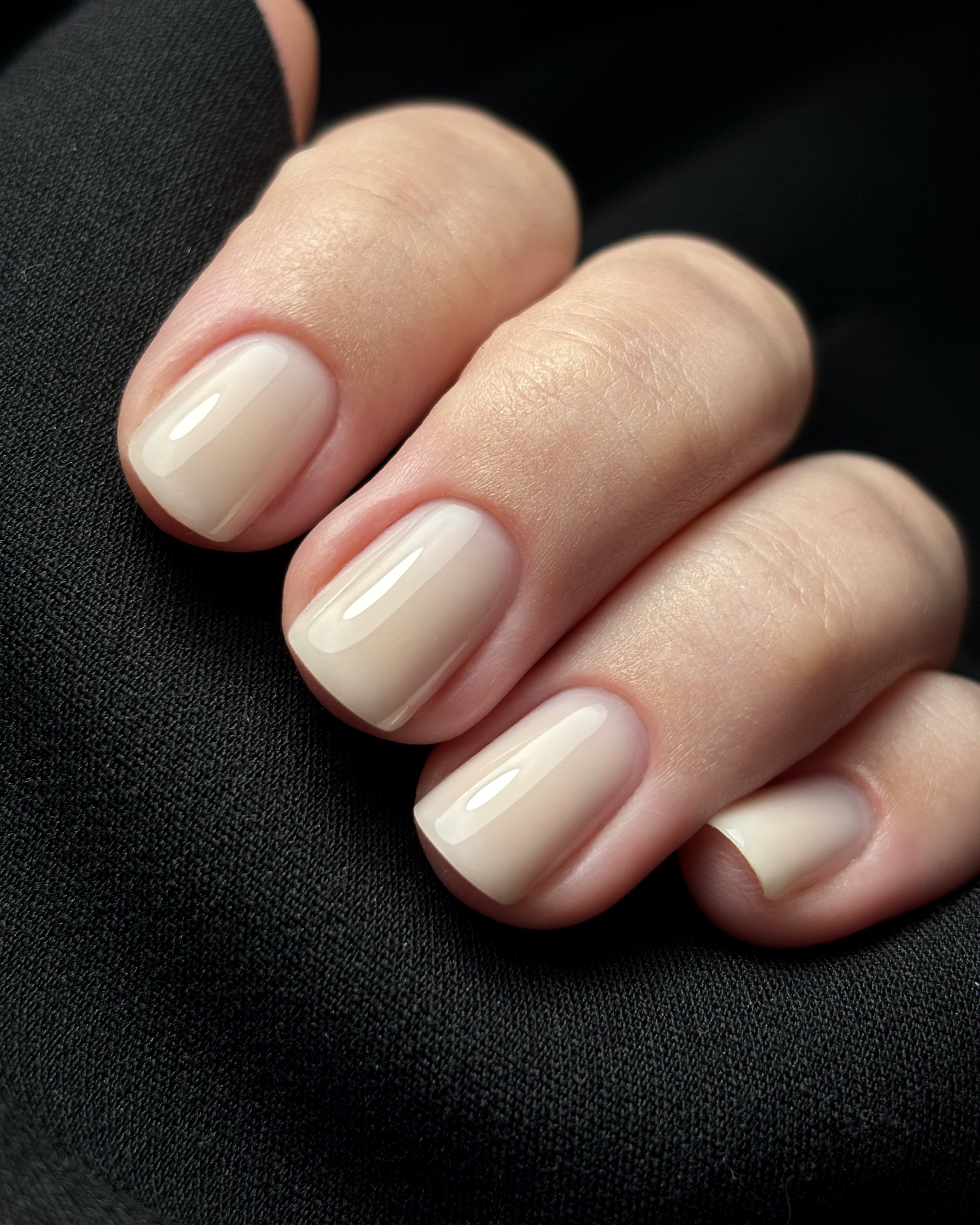 Close-up of a person's well-manicured nails with a clear or pale pink gloss, resting on black fabric.