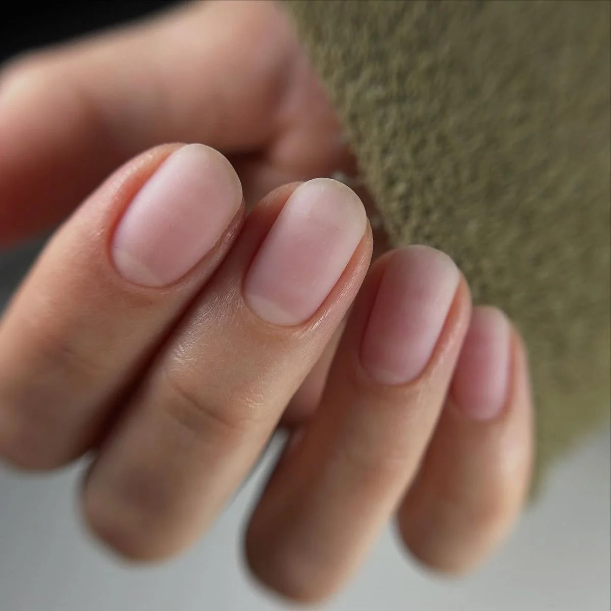 Close-up of four neatly trimmed fingernails with natural nail color, resting gently against a textured gray surface.