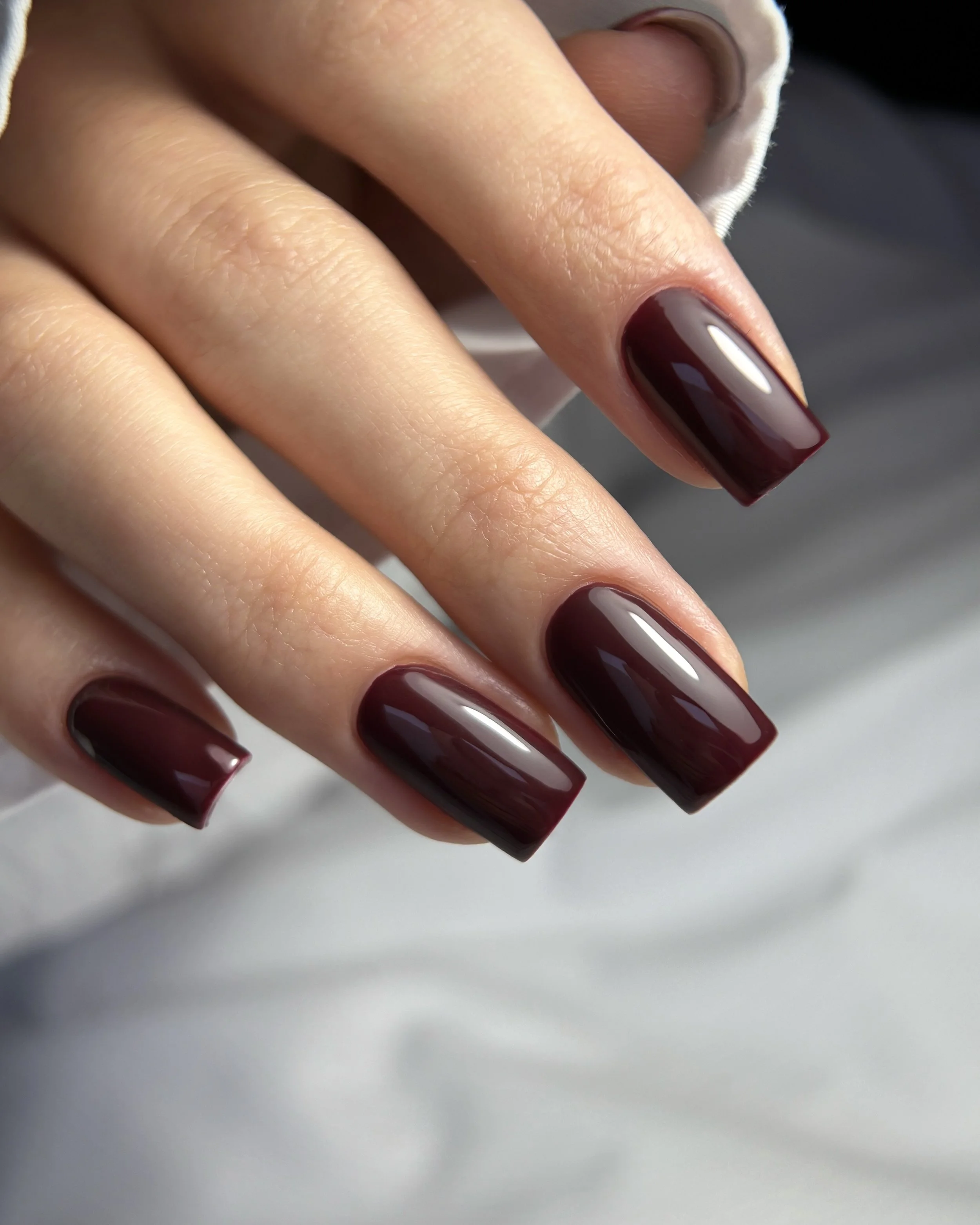 Close-up of a hand with manicured nails painted in dark burgundy, resting on a white surface.