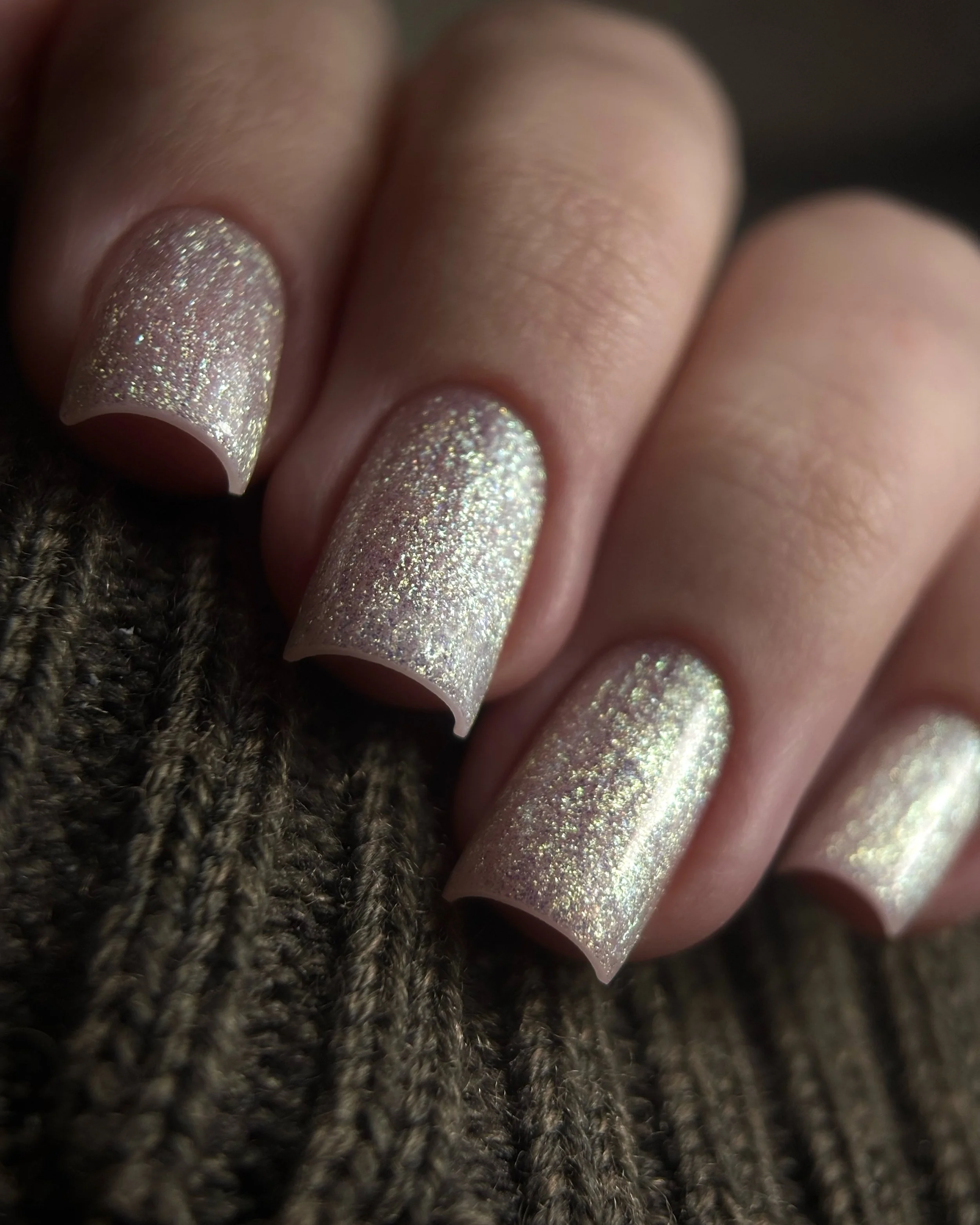 Close-up of manicured fingernails with glittery, iridescent nail polish, resting on a textured knitted fabric.