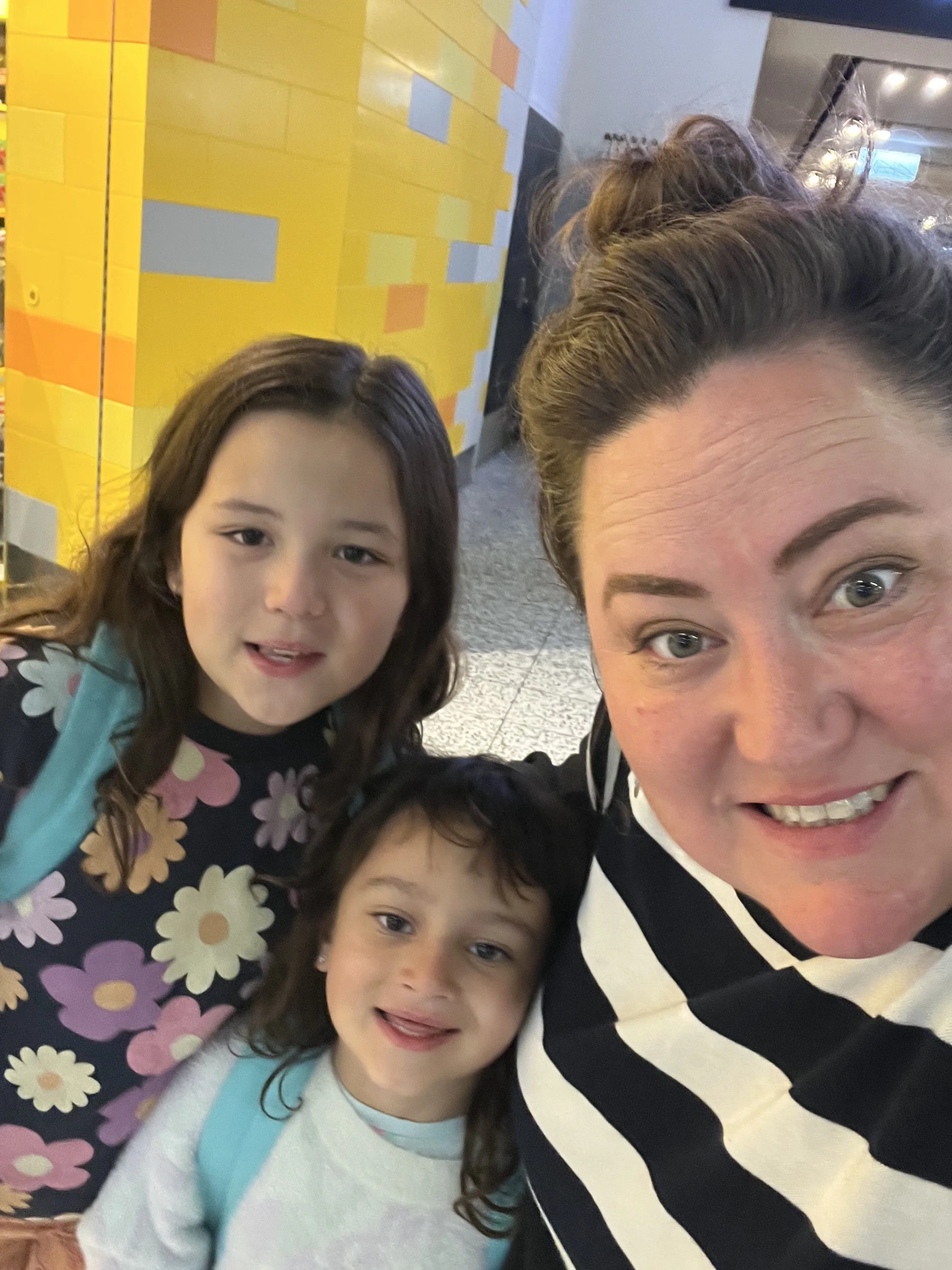 Valentina, founder of Whimsy Disco, smiling with her two young daughters in a close-up indoor selfie with a colourful background.