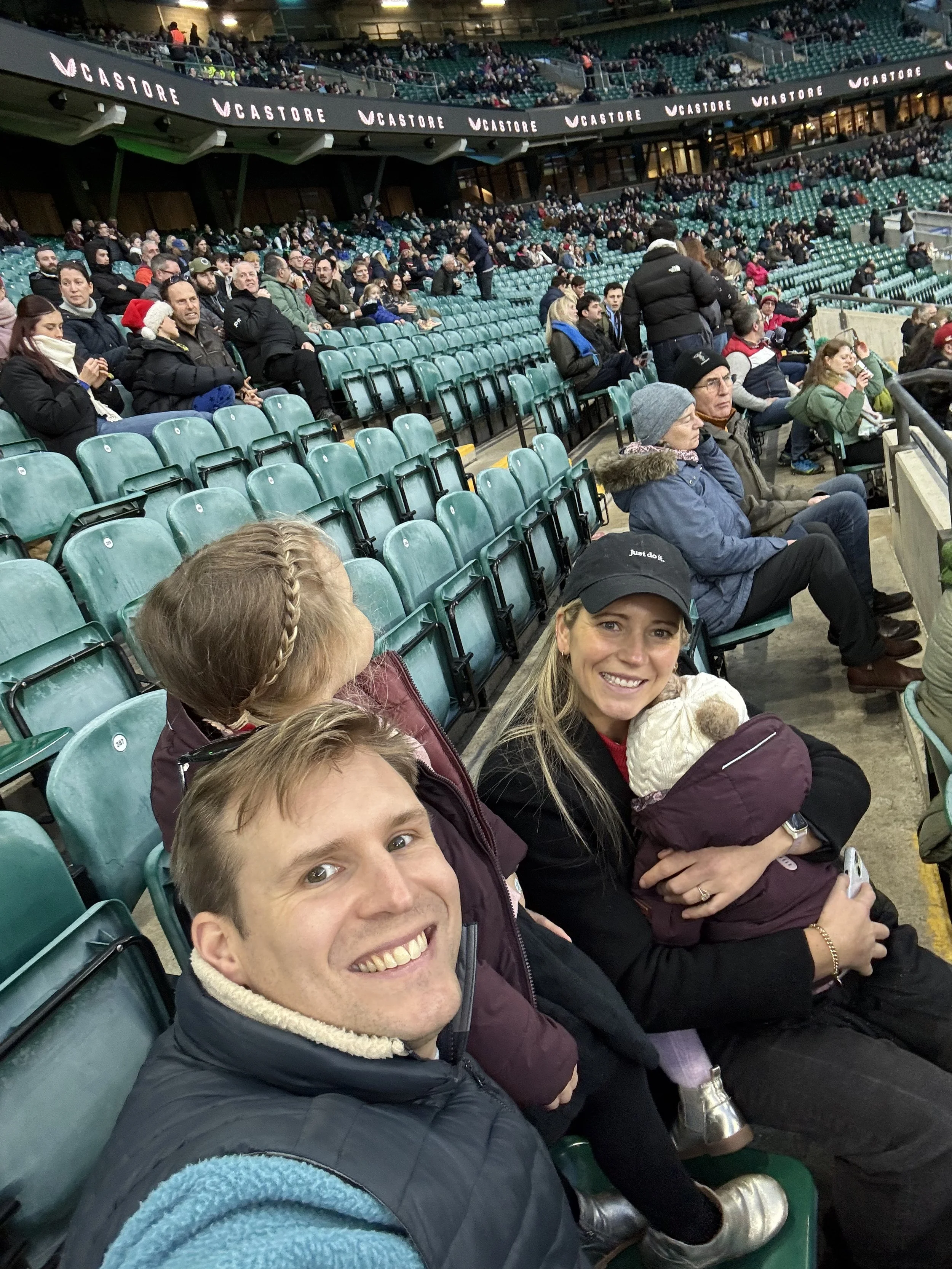 Smiling group of people, including children and adults, sitting in stadium seats during an event, with some wearing winter clothing and hats.