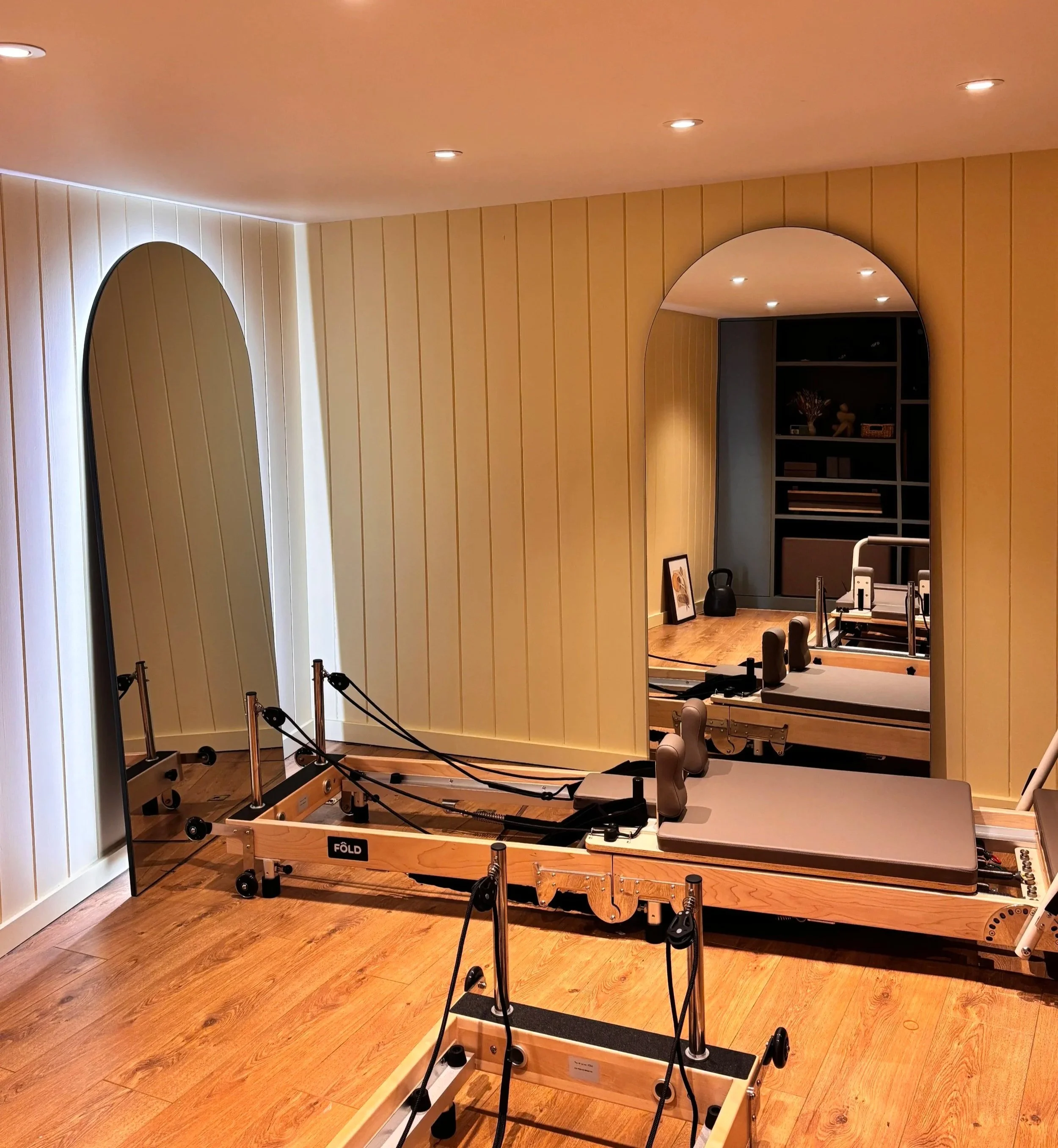 A room with wooden flooring and beige walls, featuring an exercise machine frame but no equipment. There is a mirror on one wall and a bookshelf with decor items in the background. Recessed ceiling lights are also visible.