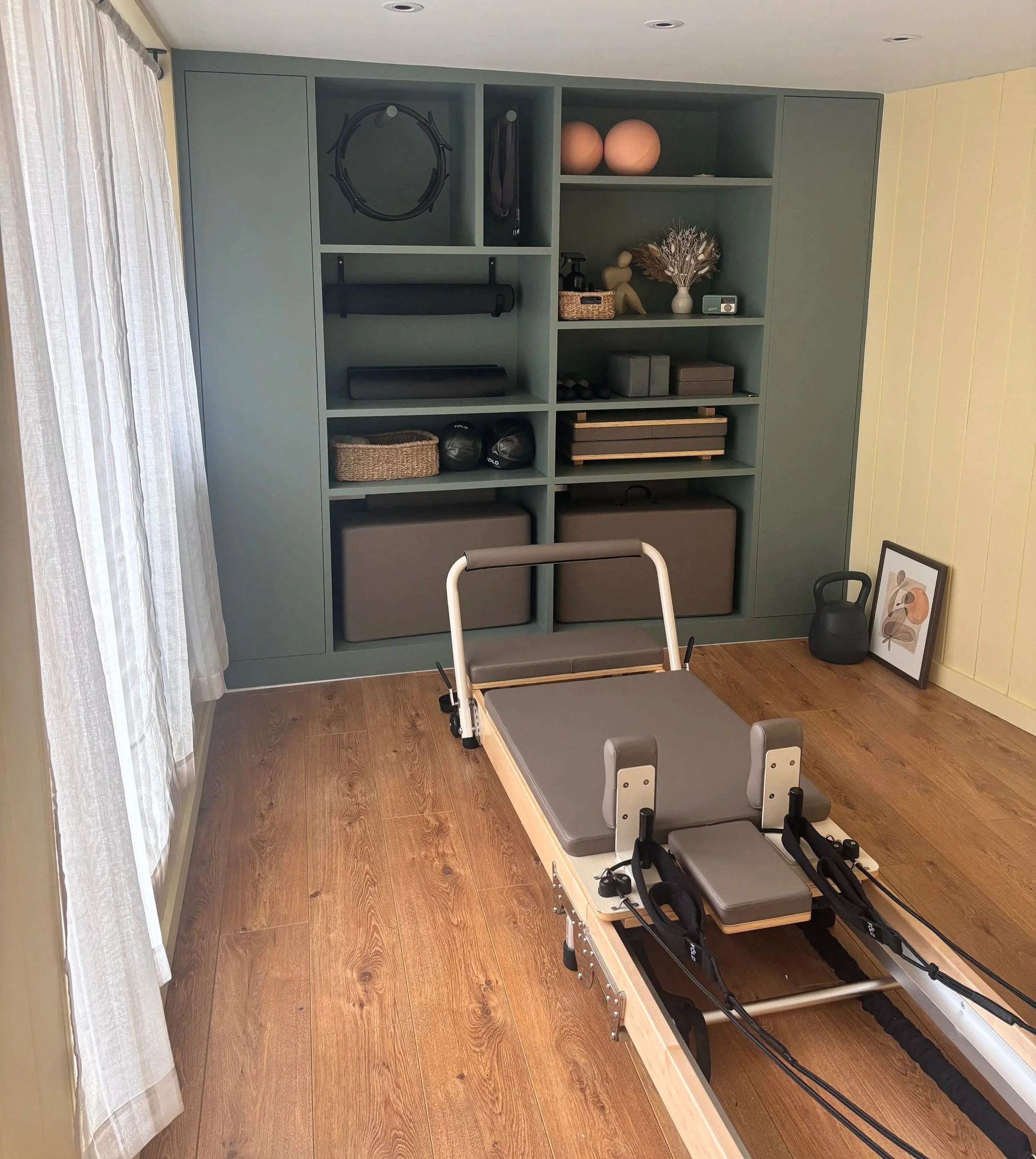 Exercise room with a green storage unit holding fitness equipment, a Pilates reformer machine with hand weights nearby, a black kettlebell, framed artwork, and window curtains.