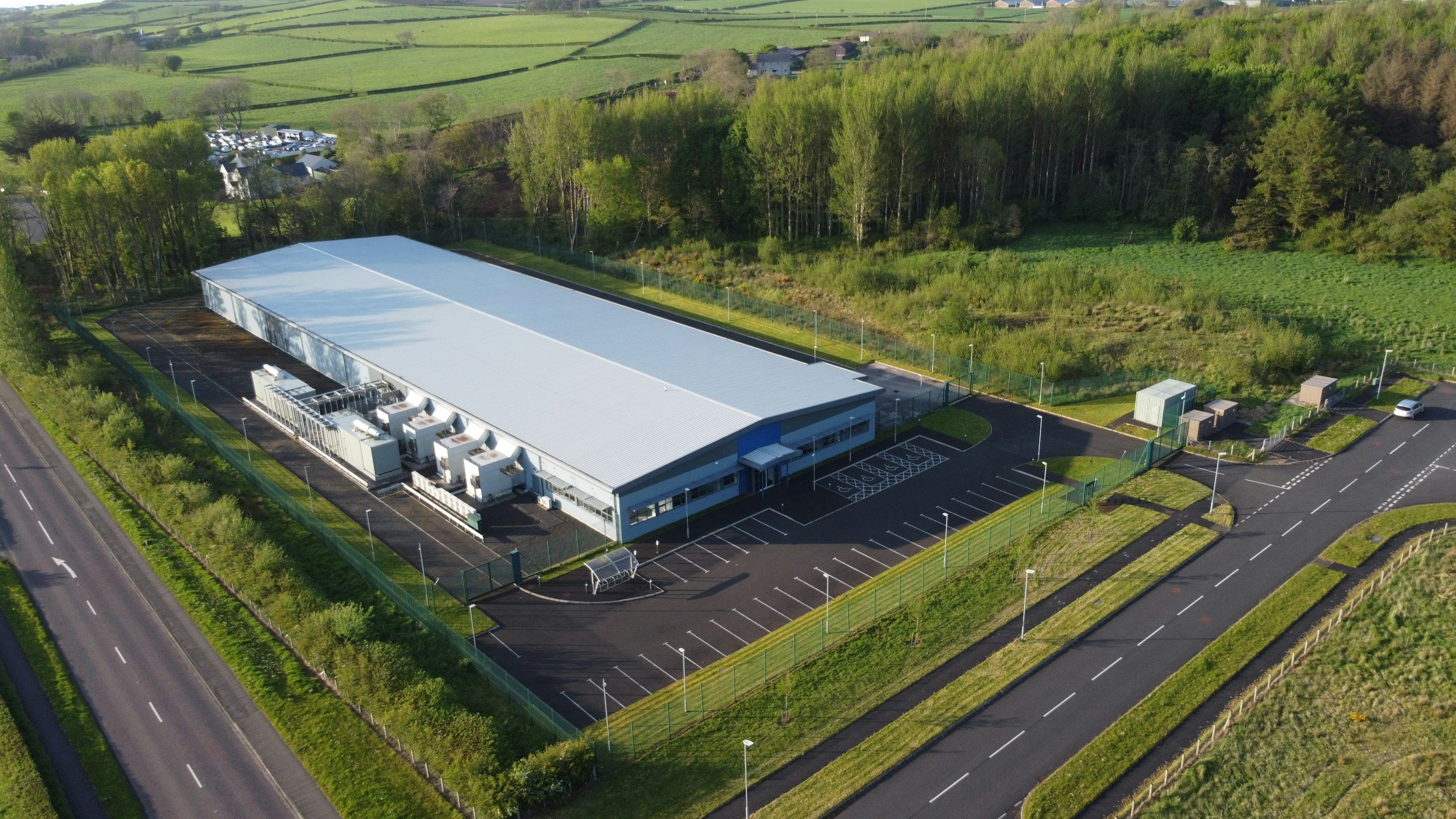 An aerial view of a large industrial building with a white roof, surrounded by parking lots, greenery, and trees, with roads nearby.