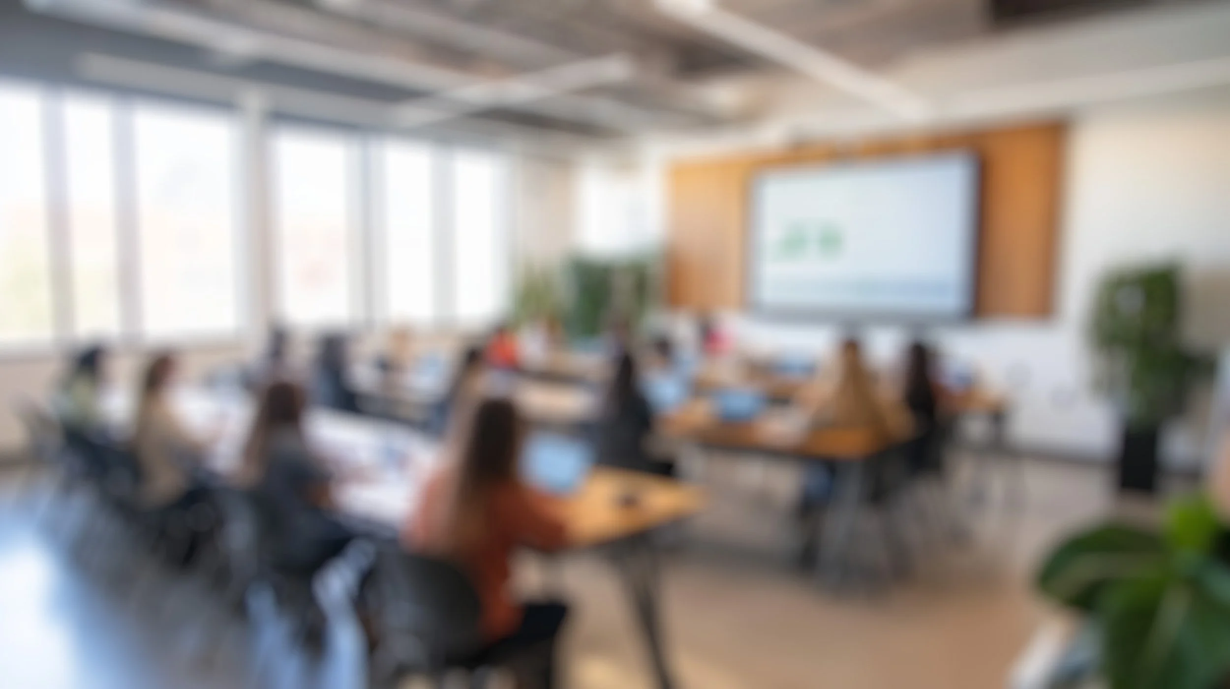 A blurred image of a conference room with people sitting at desks, facing a large screen at the front of the room.