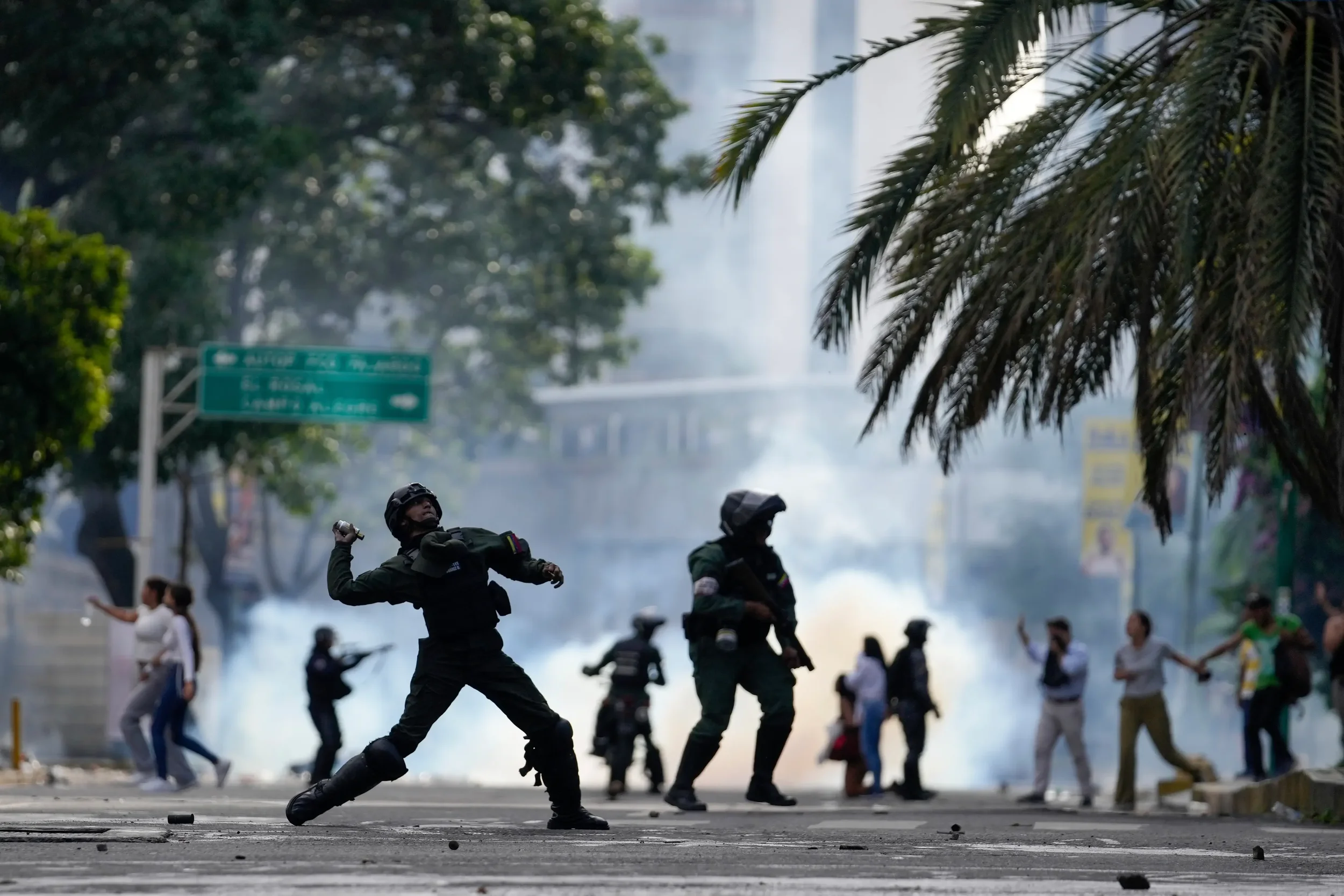 Police officers using shields and batons during a protest or riot with tear gas or smoke in the background, and protesters on the street.