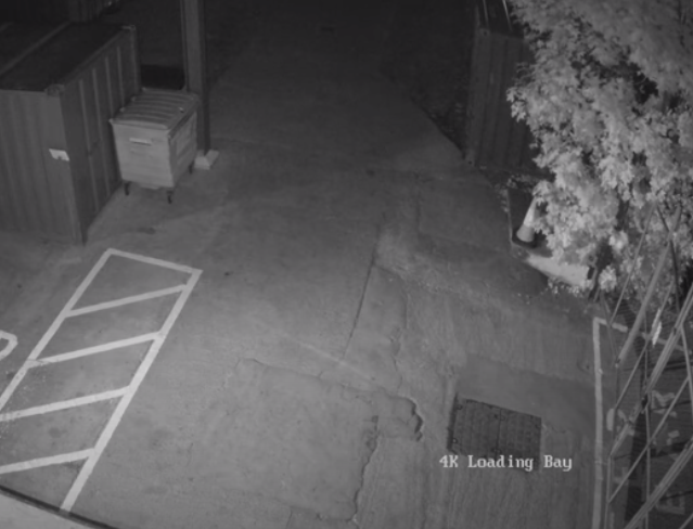 Black and white night vision camera view of a parking lot with a marked parking space, a small building or shed, a trash bin, a tree in the top right corner, and text that reads "4K Loading Bay".