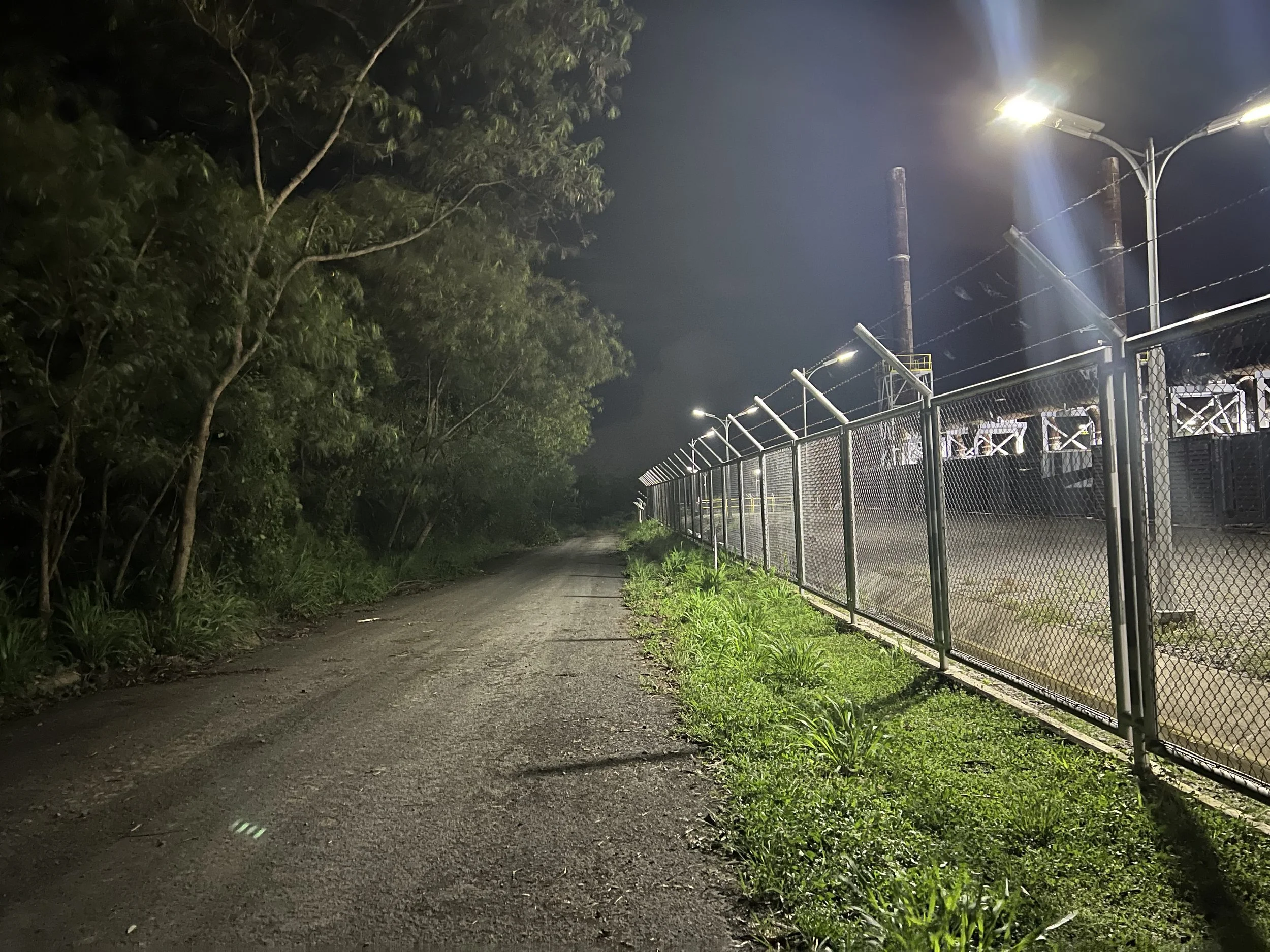 Nighttime scene of a dirt road next to a chain-link fence with barbed wire on top, lit by streetlights, with trees and industrial structures in the background.