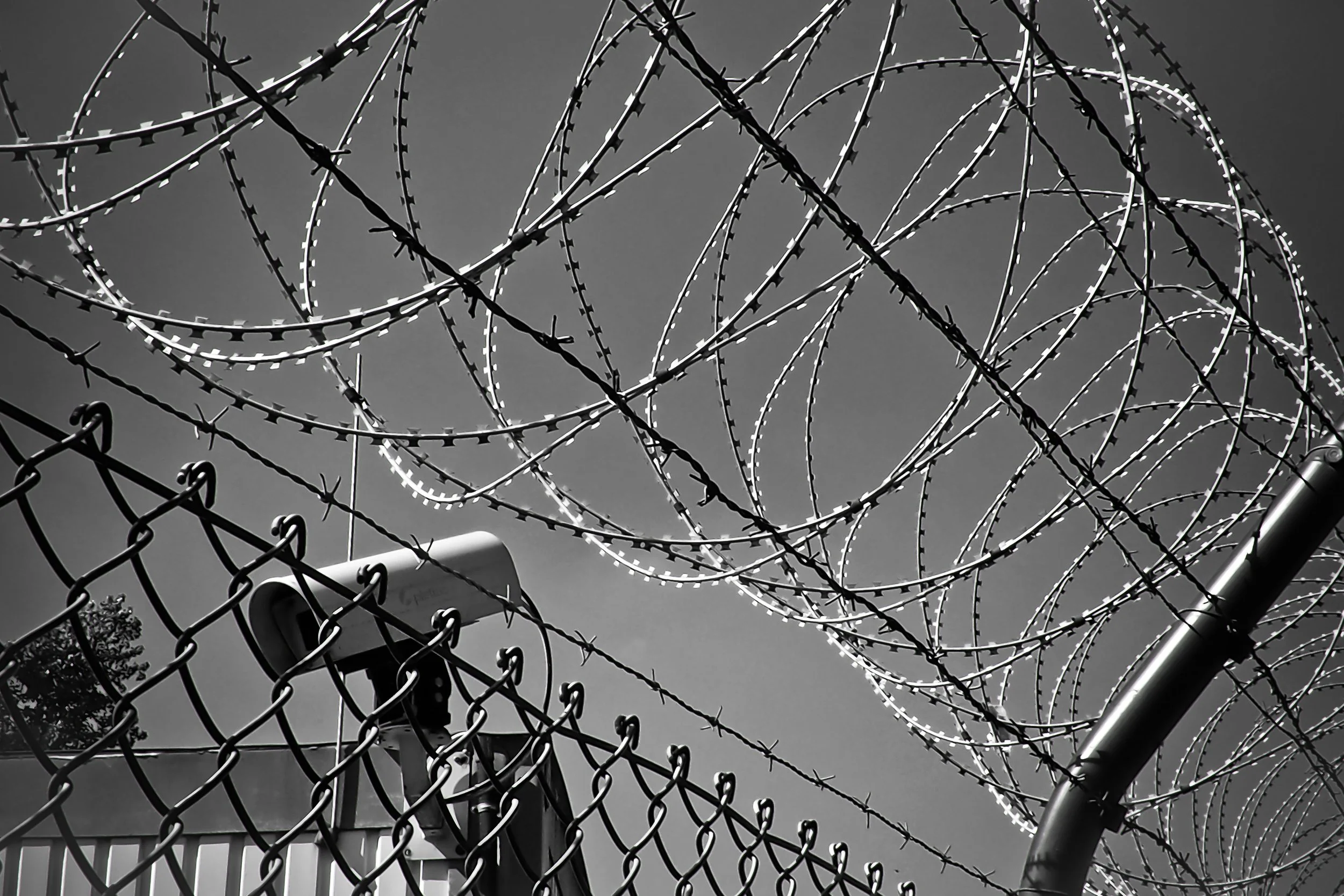 Barbed wire security fence with circular razor wire coils on top, topped with a surveillance camera, under a cloudy sky.