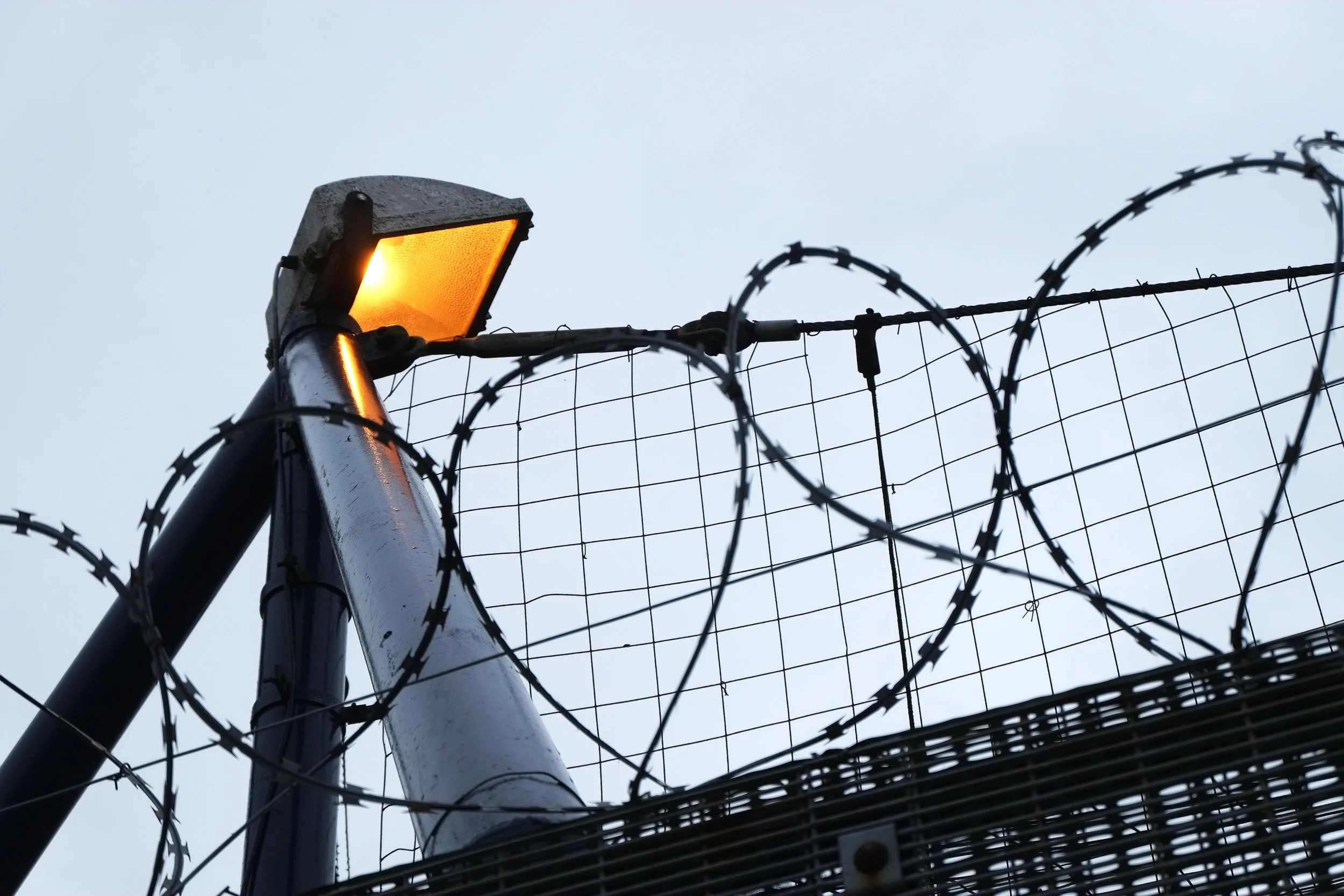 A security fence topped with barbed wire and an outdoor light fixture during daytime.