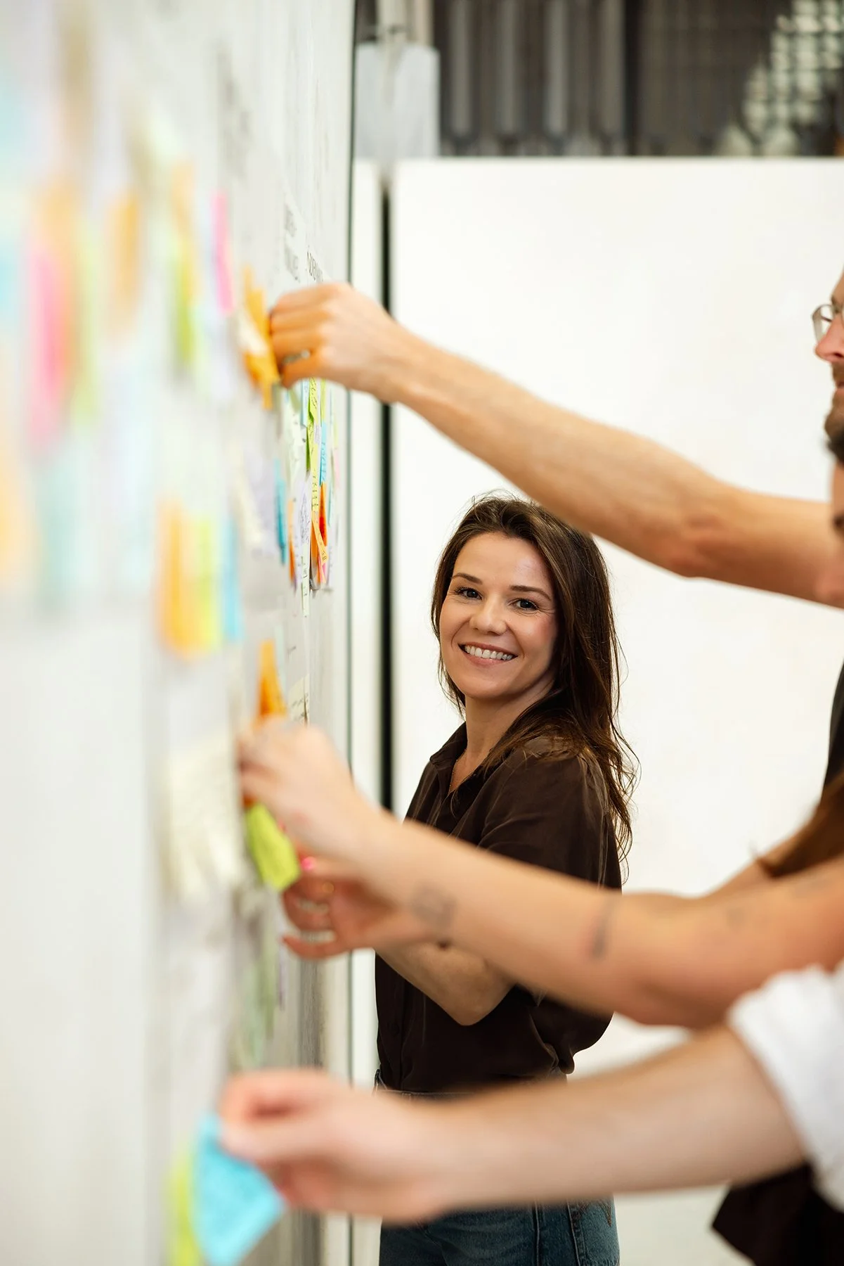 A woman smiling as she sticks notes on a whiteboard, surrounded by other people participating in a collaborative activity.