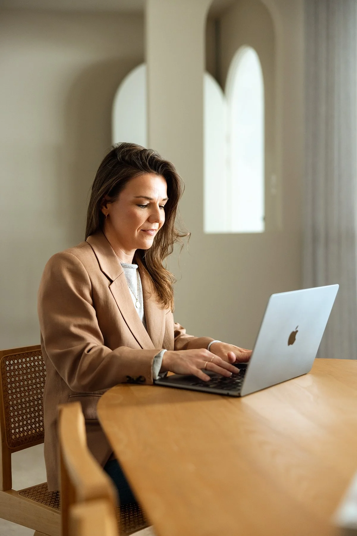 A woman with brown hair wearing a beige blazer sitting at a wooden table, working on a laptop in a room with arched windows and neutral-colored walls.