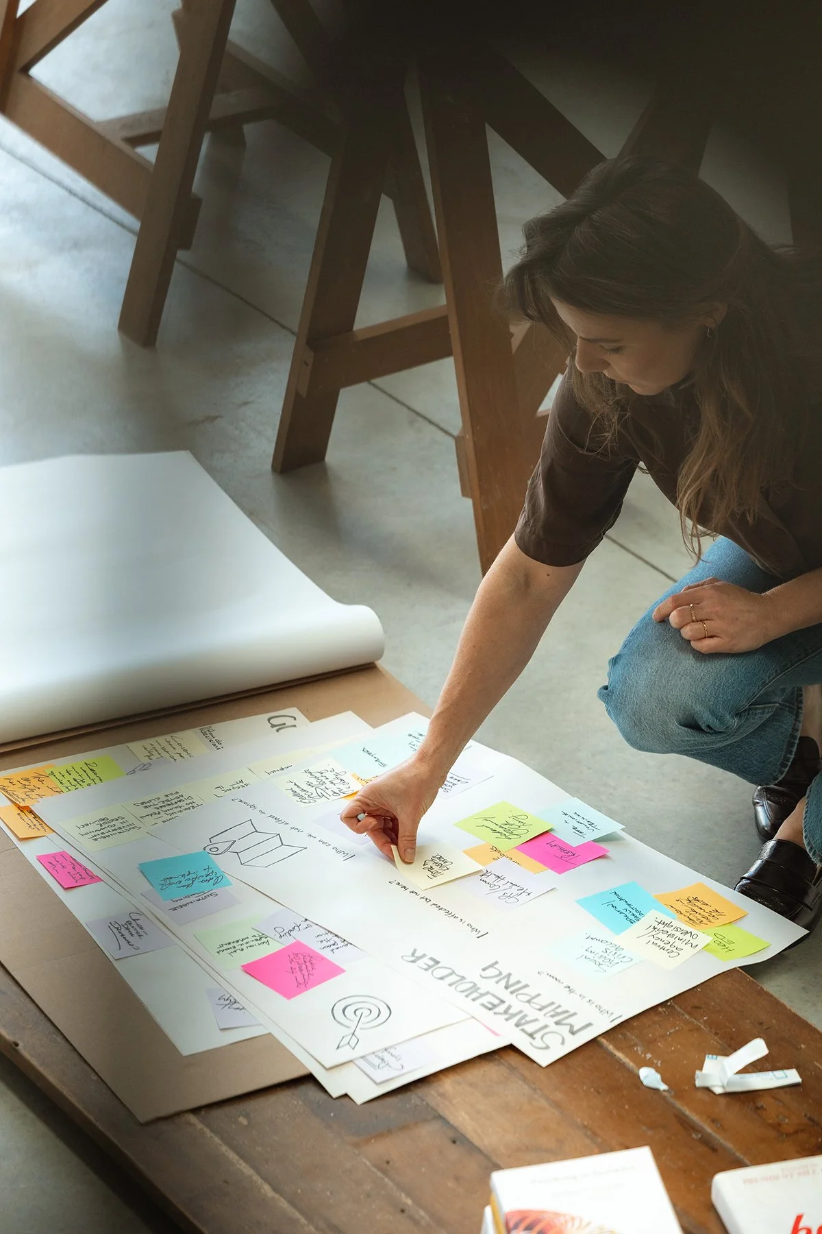 Woman in brown shirt and jeans is organizing colorful sticky notes and sketches on a large sheet of paper on a wooden table, with multiple chairs around.