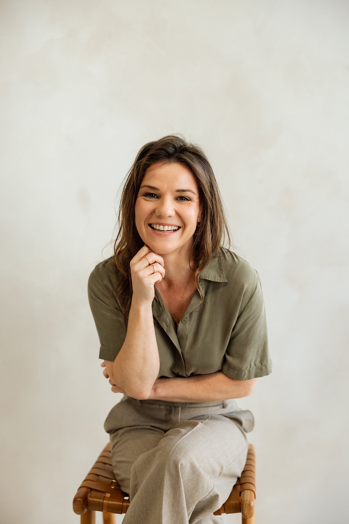 A smiling woman with brown hair, wearing a beige button-up shirt and khaki pants, sitting on a wooden stool against a plain light-colored wall.