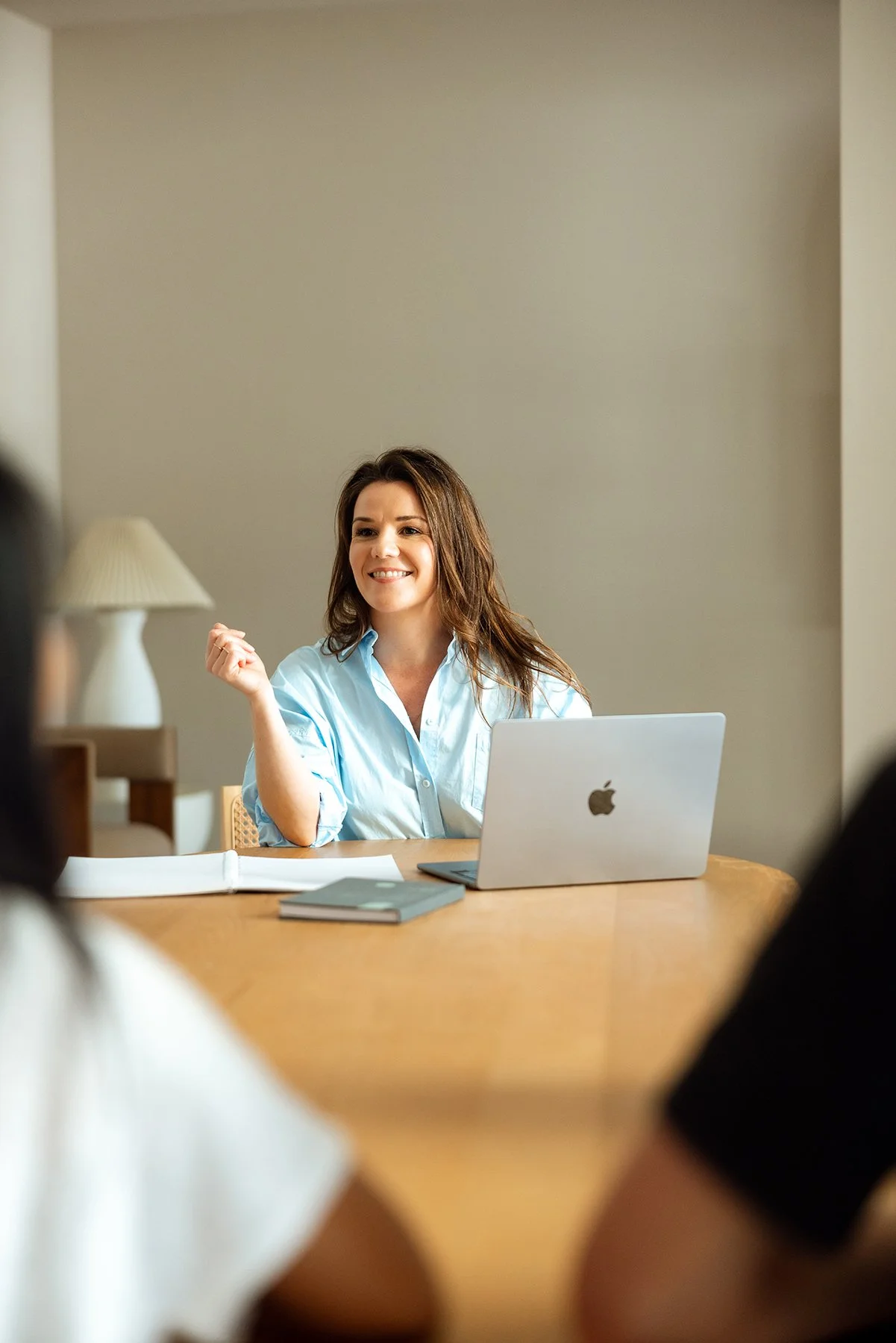 A woman with brown hair and a light blue shirt smiling during a meeting at a wooden table with an open laptop, notebooks, and papers. The background is a plain beige wall with a white lamp visible.