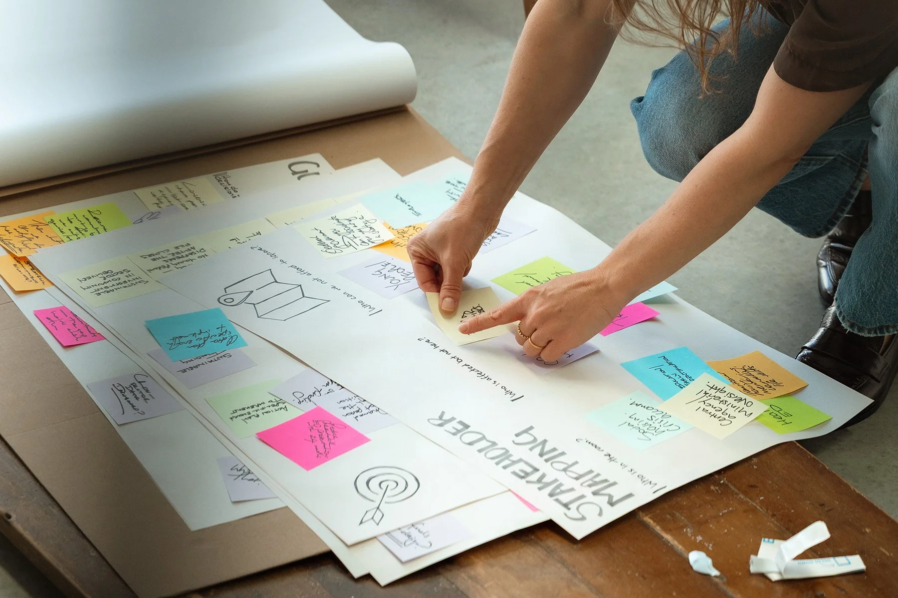 A person arranging colorful sticky notes on a large paper with handwritten notes and diagrams, on a wooden table.