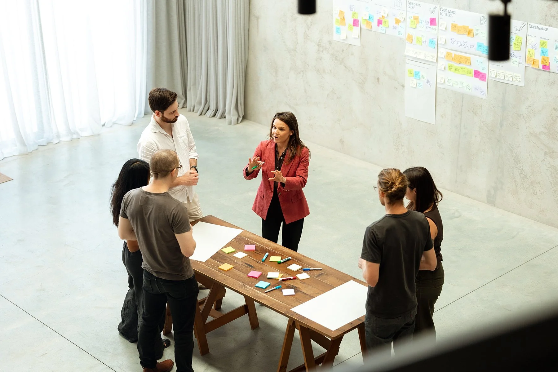 A woman in a red blazer leads a team meeting in a bright conference room with large windows and white curtains. The group of five mixed-gender people listens attentively, standing around a wooden table with colored sticky notes and markers. On the wall behind them, there are large sheets of paper with various notes and diagrams.