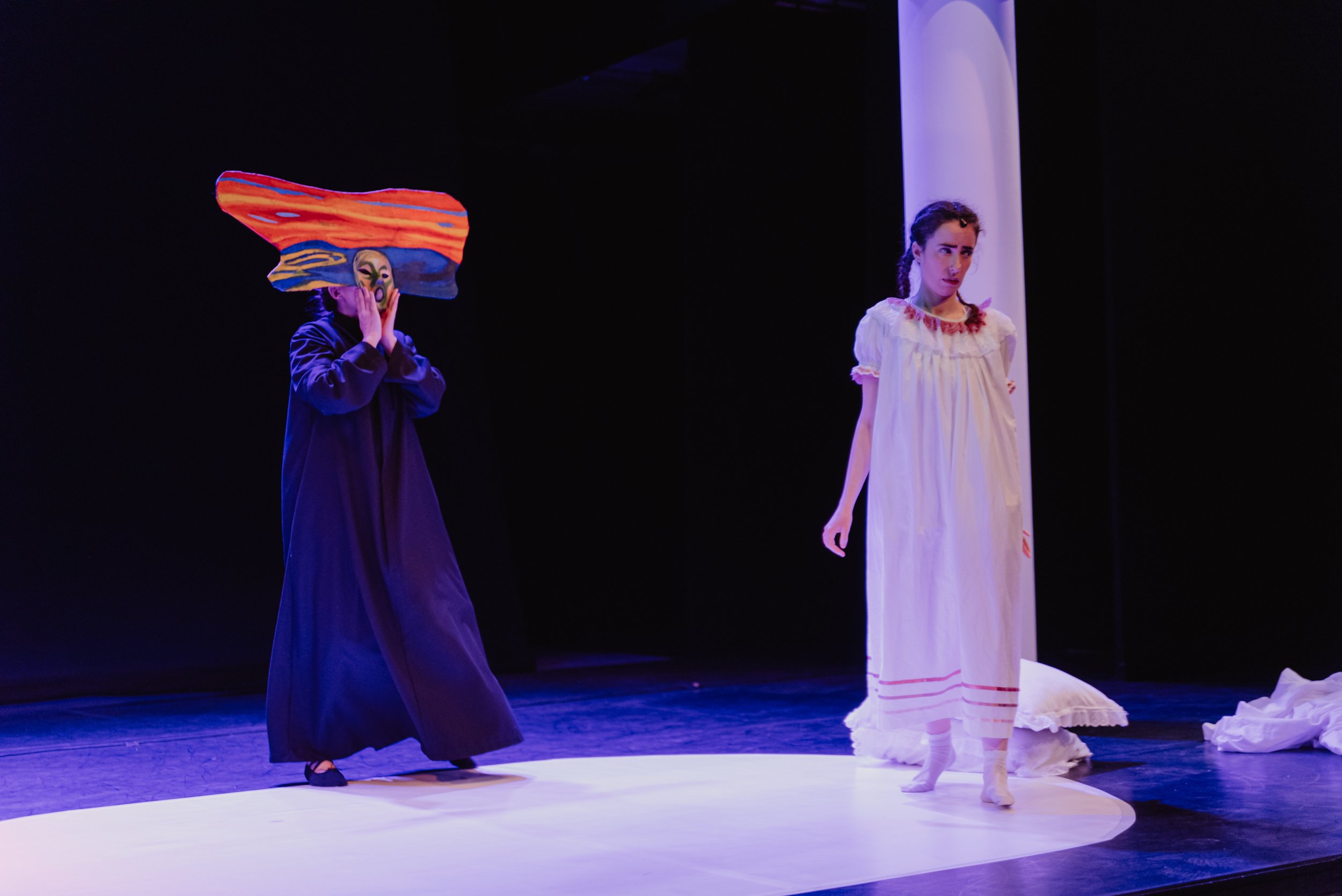 Dos mujeres en un escenario teatral, una con máscara y cabeza de pintura abstracta, la otra con vestido blanco, en medio de una puesta en escena artística.