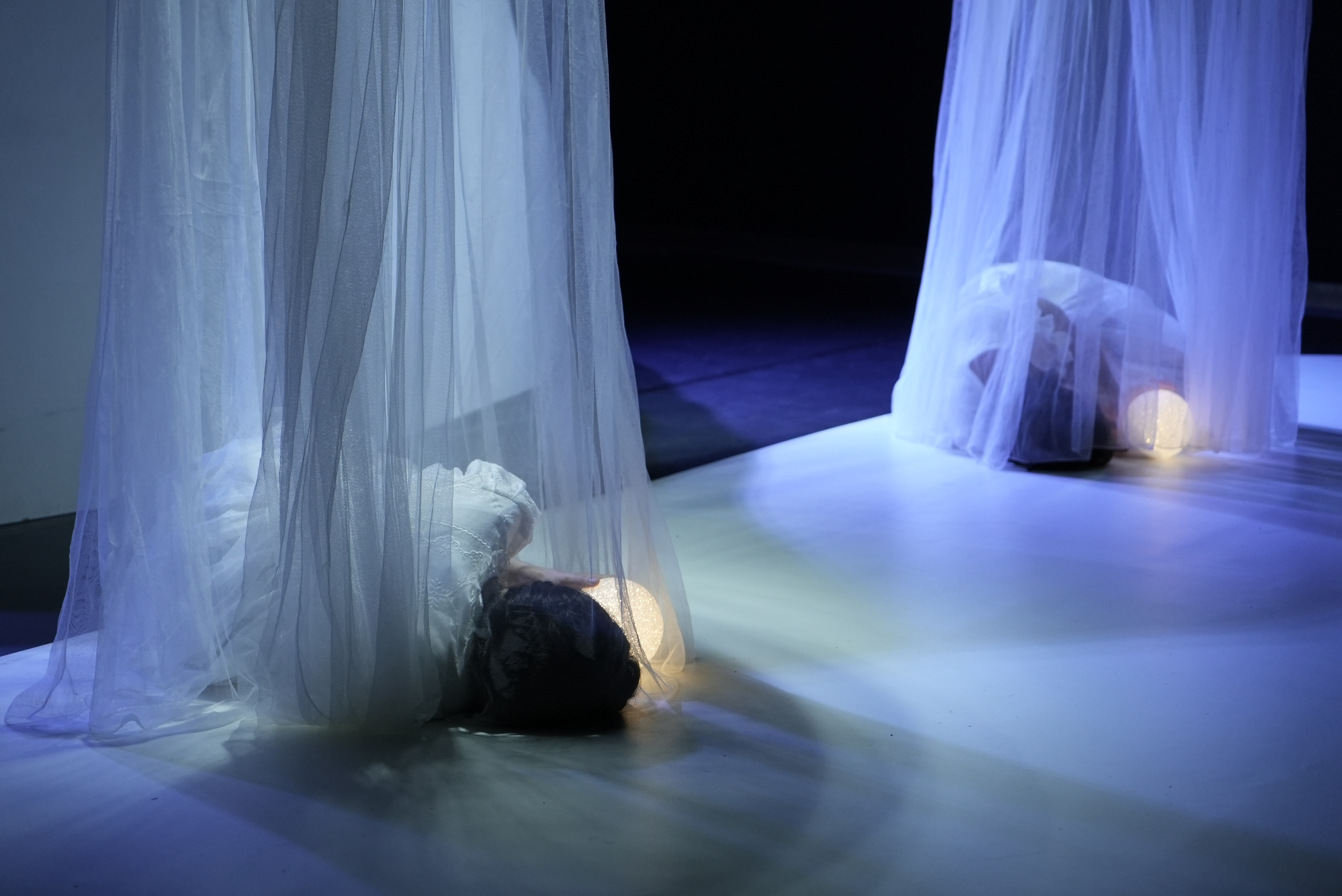Persona acostada en el suelo, cubierta con tela blanca, vista a través de una cortina de tela similar, en un entorno oscuro con iluminación suave.